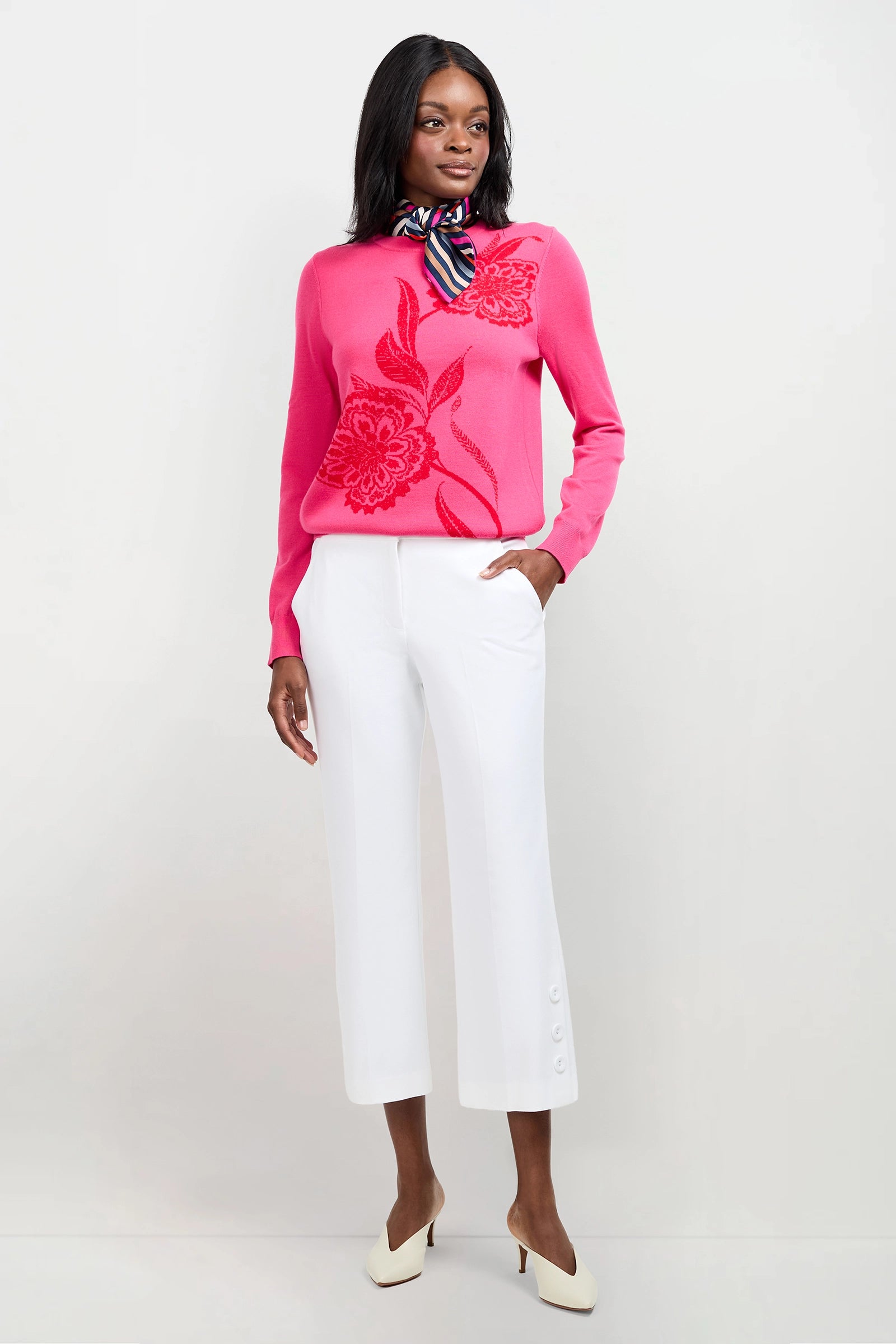 A woman stands against a plain background wearing the Old Fashioned Sweater, a bright pink top with a large floral pattern, paired with white cropped pants with button details, white heels, and a colorful striped scarf around her neck.