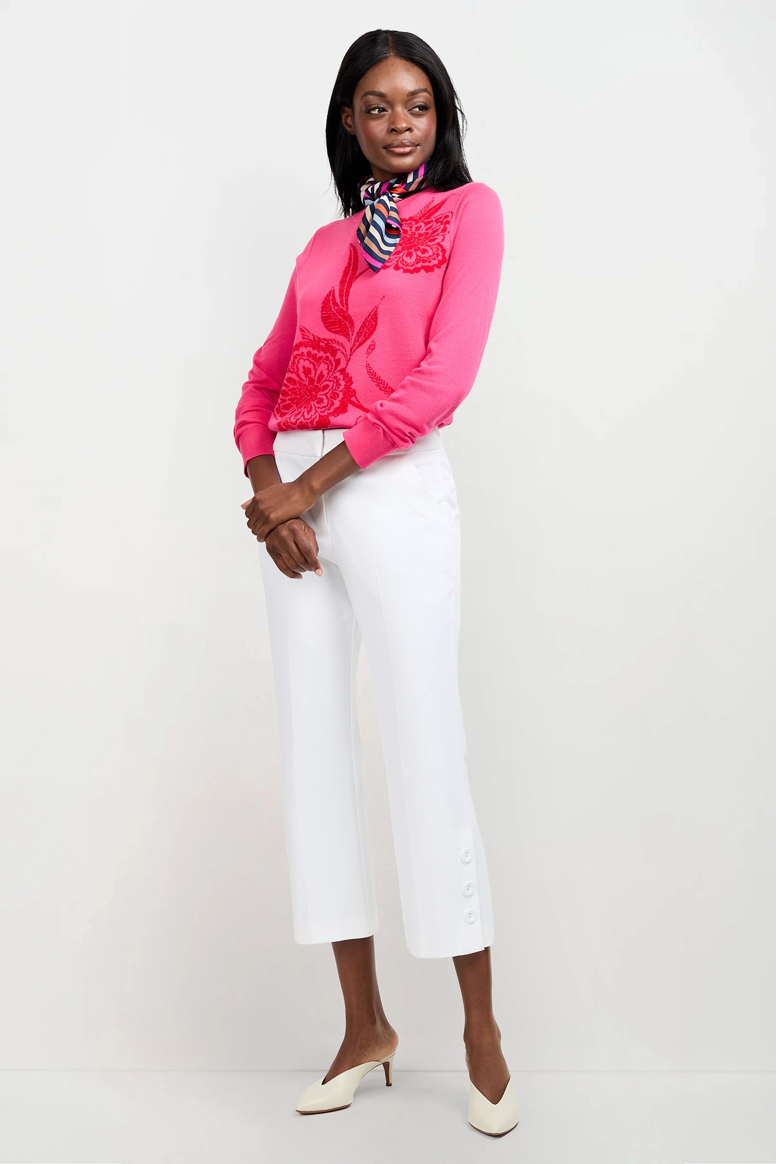 A woman poses against a white background wearing the Old Fashioned Sweater in bright pink with a floral pattern, paired with white cropped pants featuring button details, beige heels, and a colorful striped scarf.