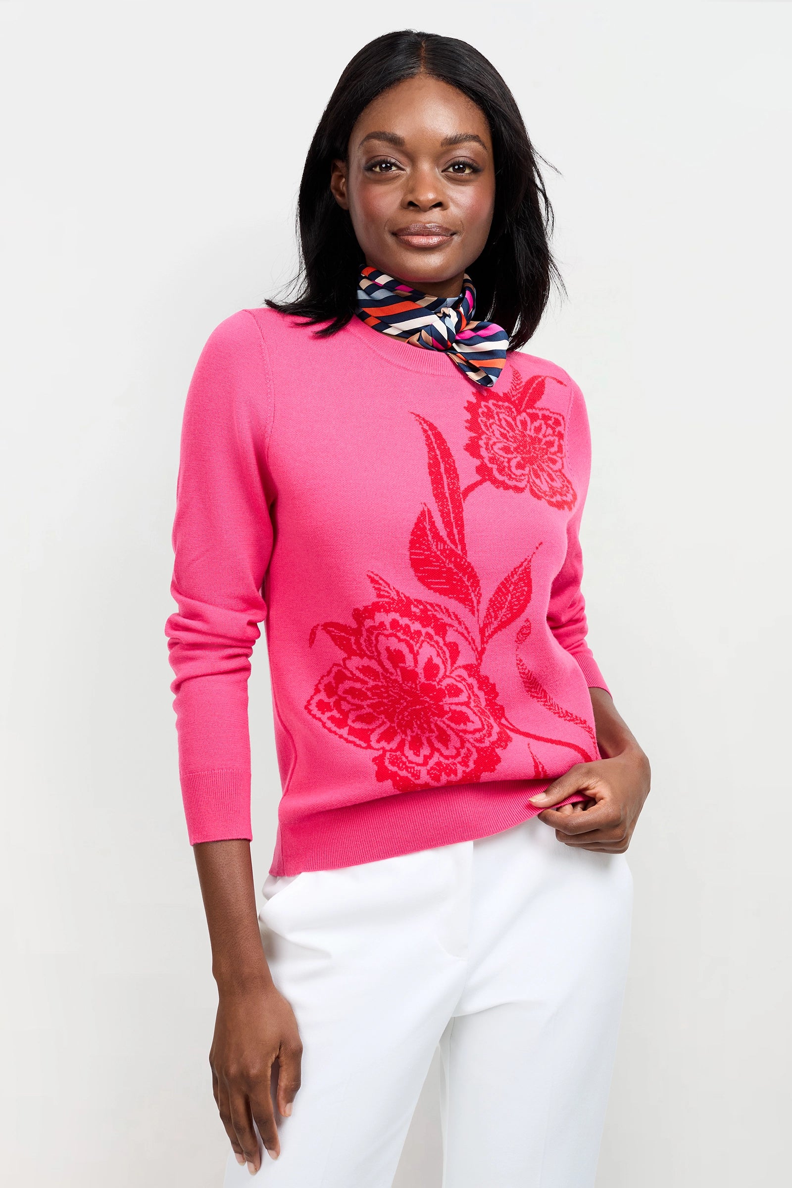 A woman smiles gently with one hand in her pocket, wearing the Old Fashioned Sweater—a bright pink top with large floral designs—paired with a colorful striped scarf and white pants, standing against a plain light background.