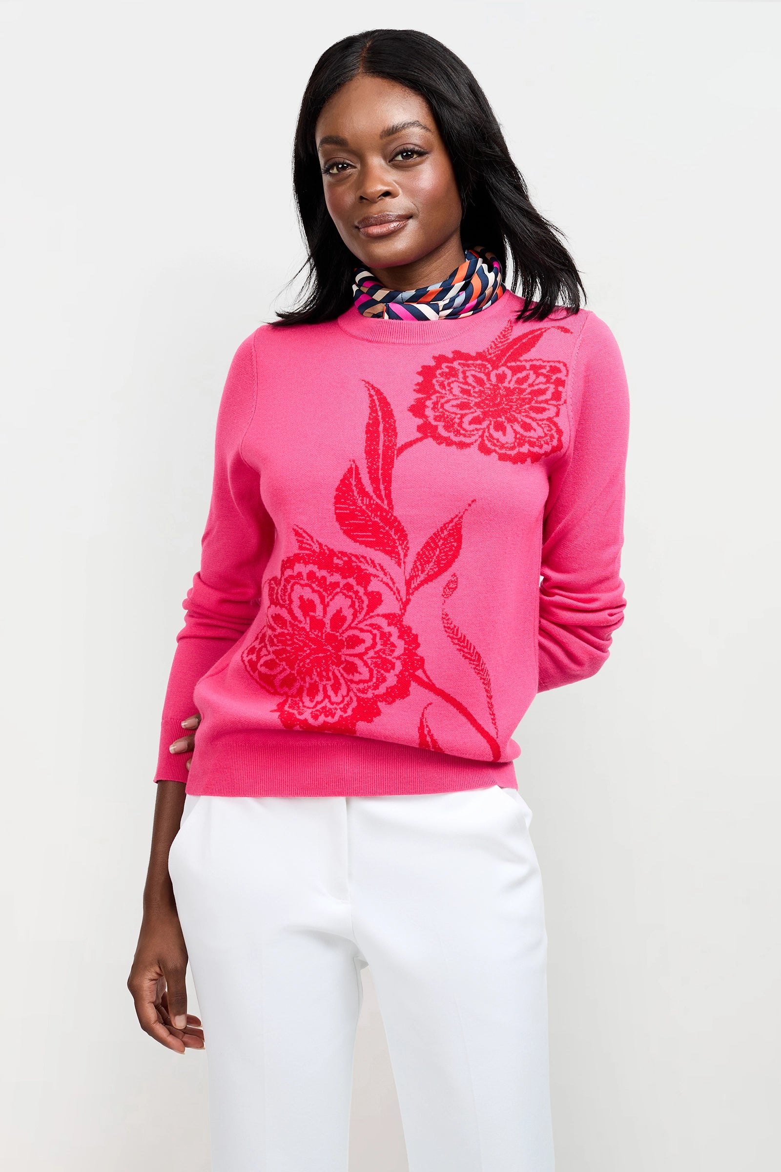 A woman smiles gently, posing against a white background in the Old Fashioned Sweater—bright pink with a red floral design—paired with white pants and a colorful patterned scarf.