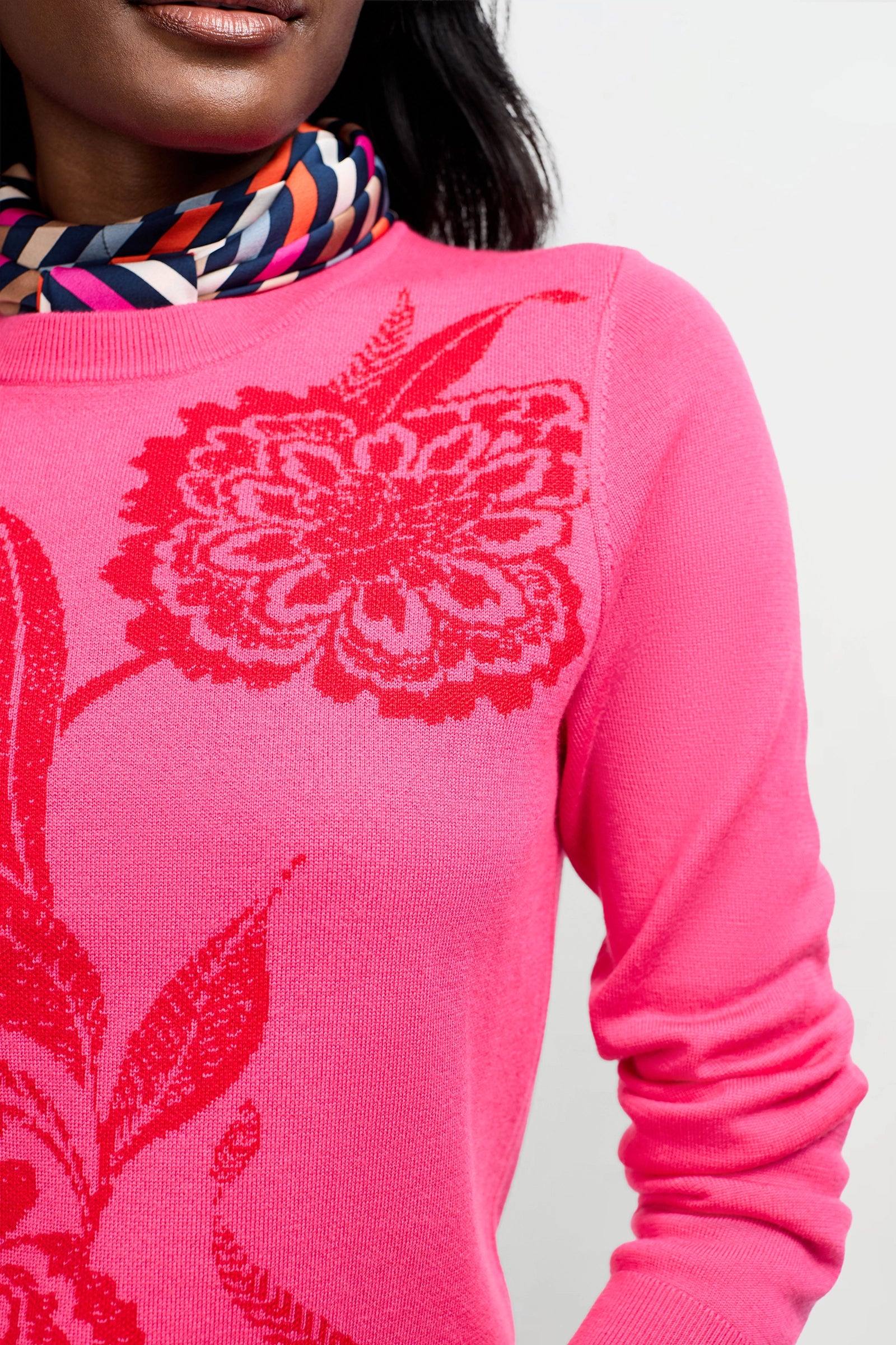 A woman wears the Old Fashioned Sweater, a bright pink top with a bold red floral pattern, paired with a colorful geometric scarf. Only her lower face, neck, and upper body are visible.
