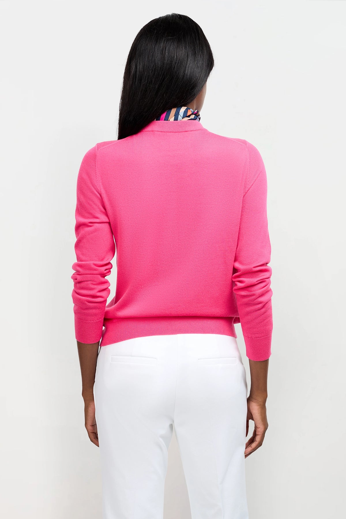 A woman with long dark hair, seen from behind, wears the Old Fashioned Sweater in bright pink and white pants against a plain light background.