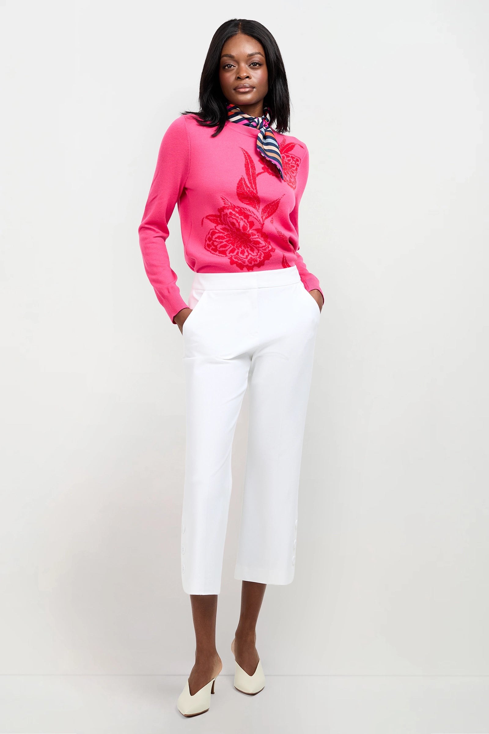 A woman stands against a white background wearing the Old Fashioned Sweater in Ferlo Floral, paired with white cropped pants, white heels, and a striped scarf. Her hands are in her pockets and she is smiling slightly.