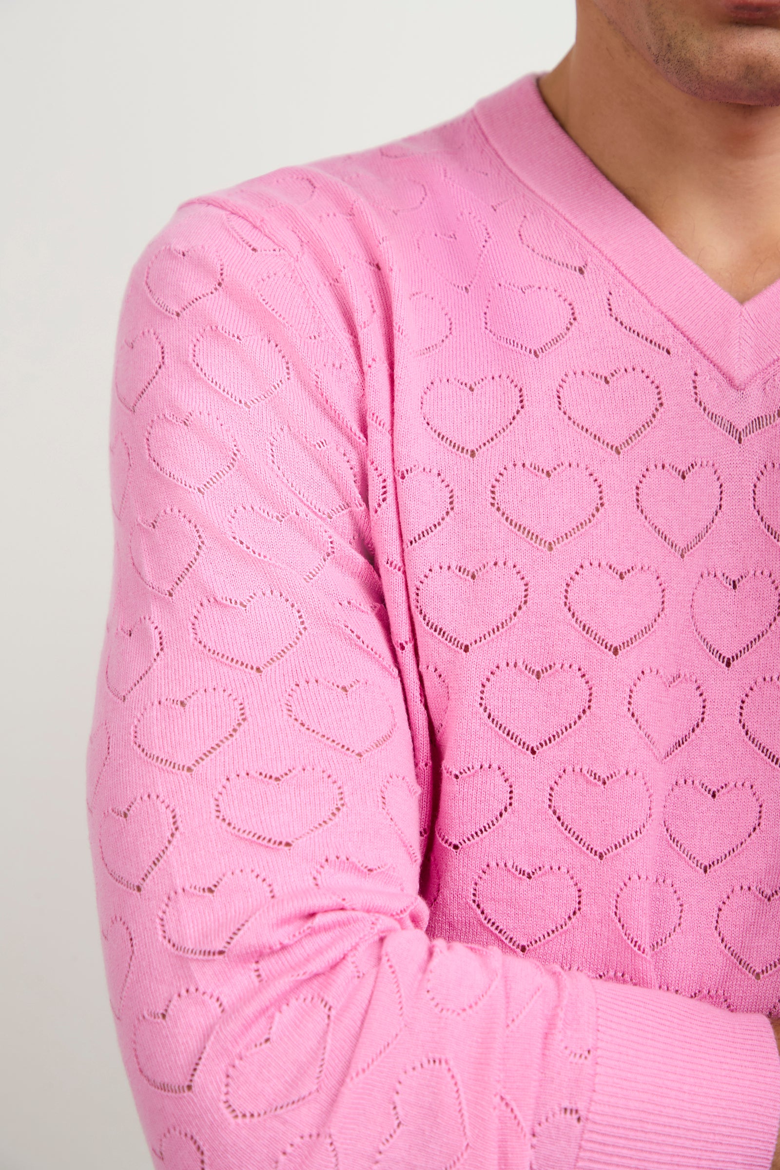 A person models the Eros Heart Pointelle V-Neck Sweater in light pink, featuring a textured pattern of small, repeating hearts. The image highlights the upper body and showcases the sweater’s heart-themed design.