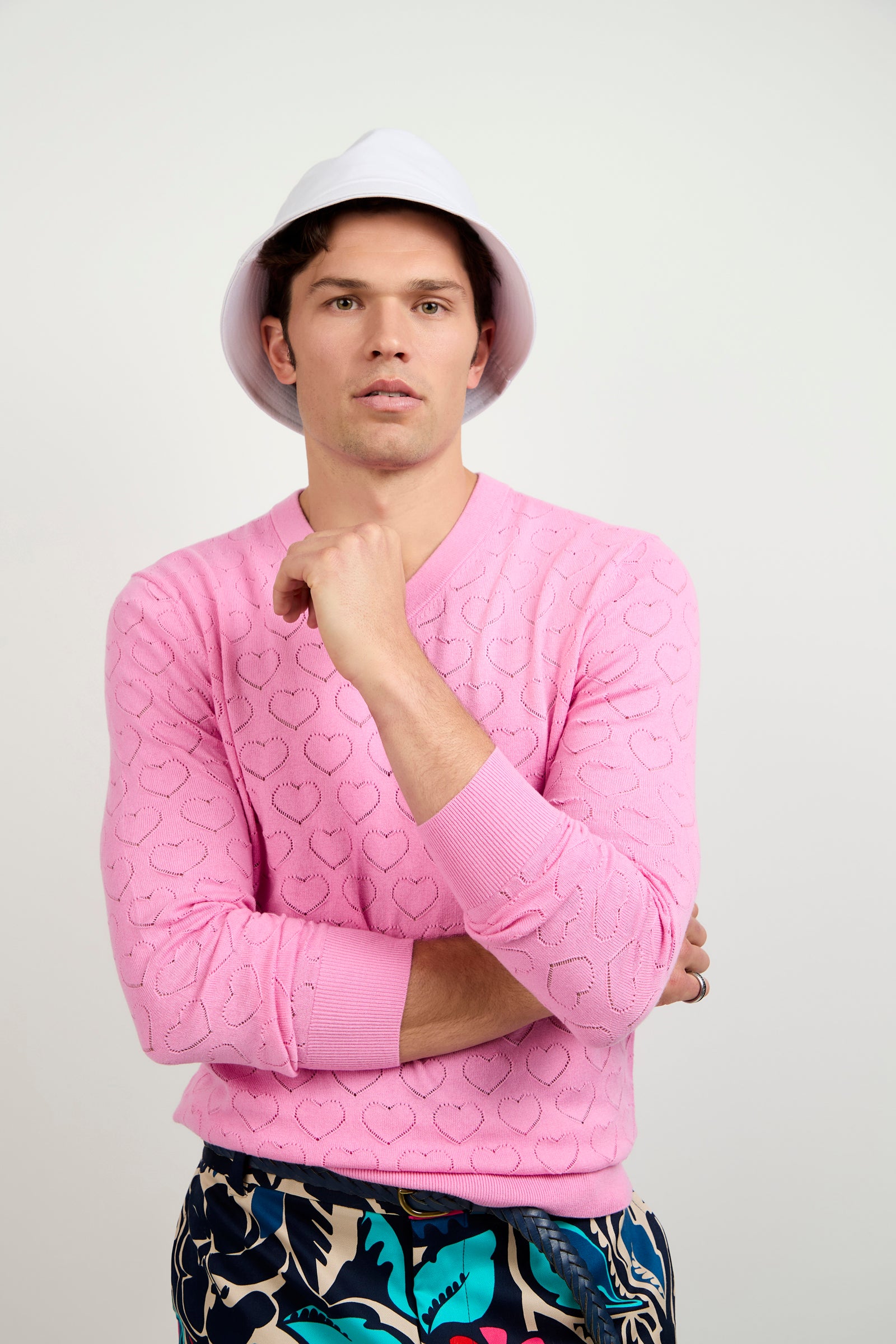 A man in an Eros Heart Pointelle V-Neck Sweater, a white bucket hat, and patterned pants stands against a plain light background with one arm crossed and the other hand near his chin.
