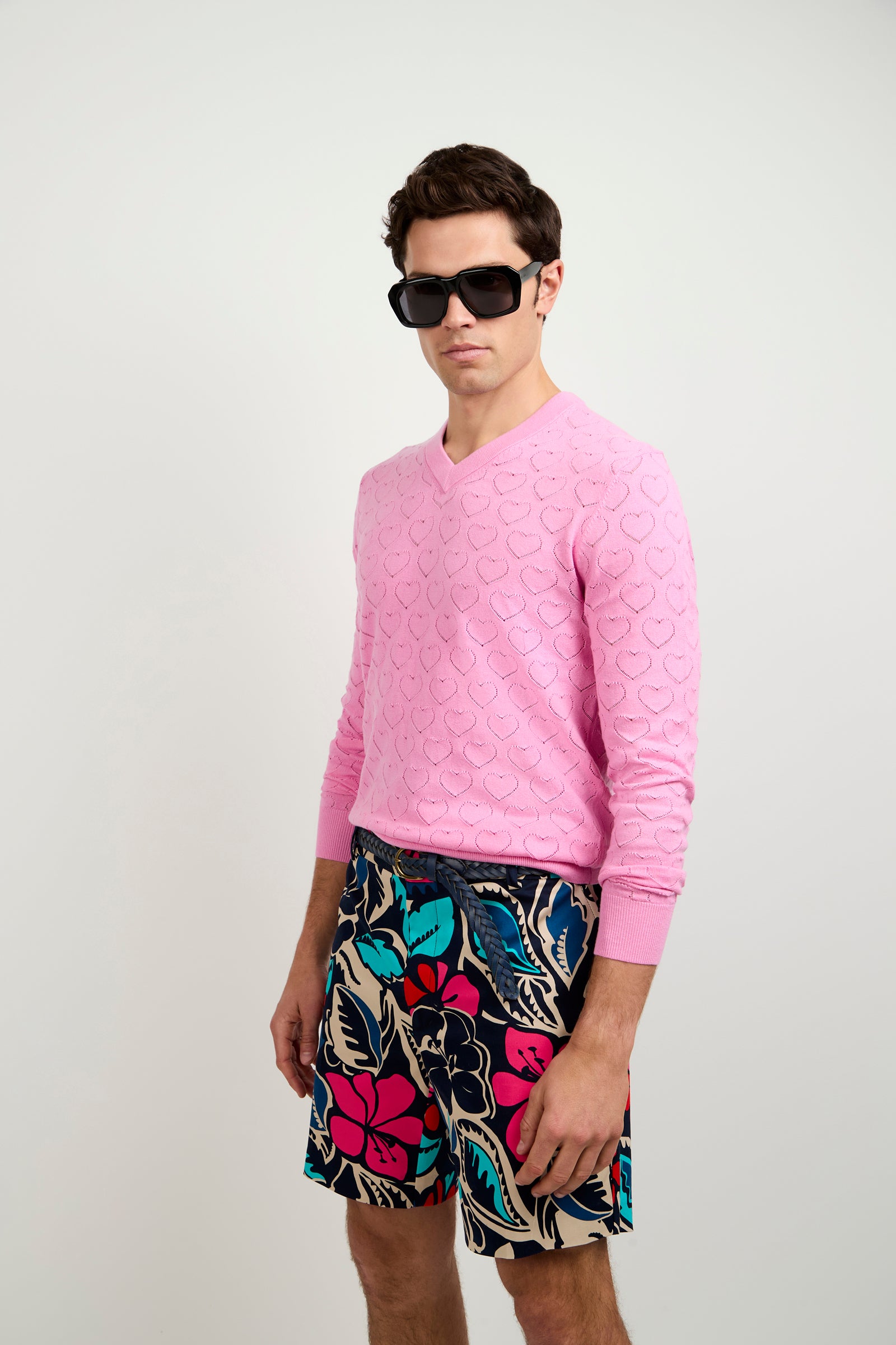 A man poses against a plain light background, wearing black sunglasses, floral shorts, and the Eros Heart Pointelle V-Neck Sweater in pink.