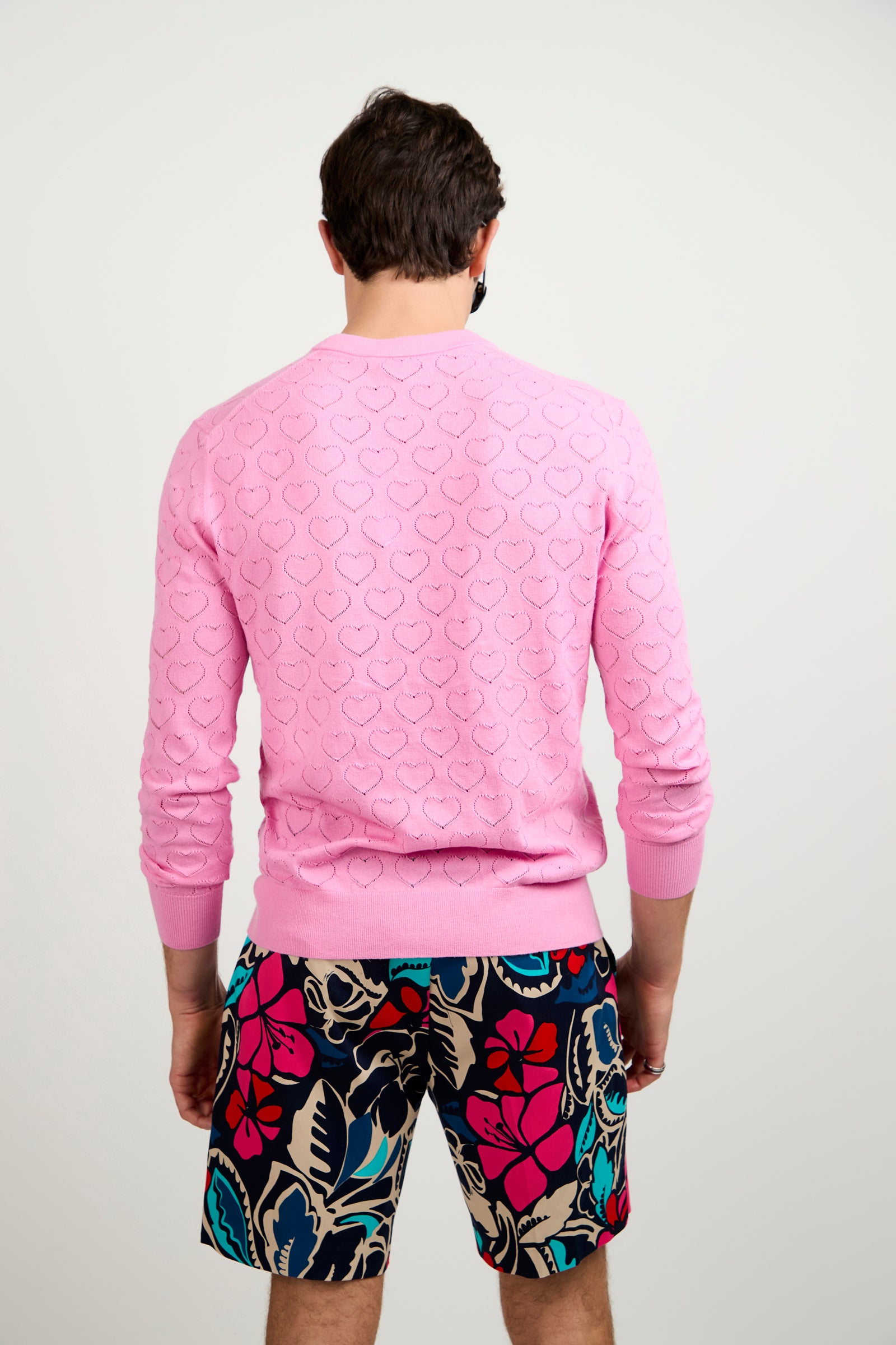 A man faces away from the camera, wearing the Eros Heart Pointelle V-Neck Sweater in pink and colorful floral shorts. The background is plain and light-colored.