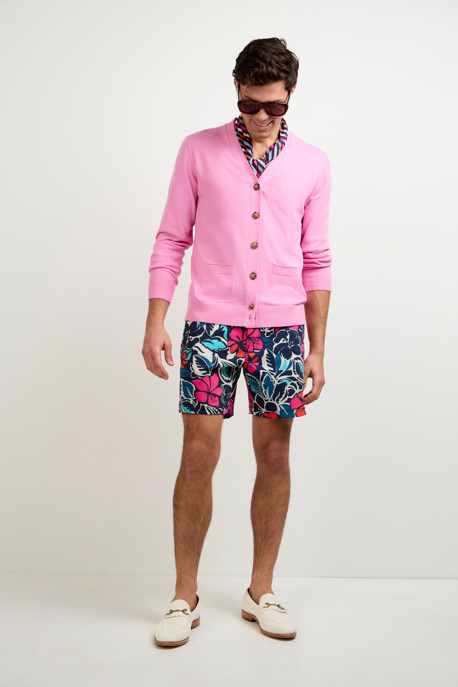A man wearing the Jacques Cardigan, floral shorts, a patterned shirt, white loafers, and sunglasses stands against a plain white background, looking down and smiling.