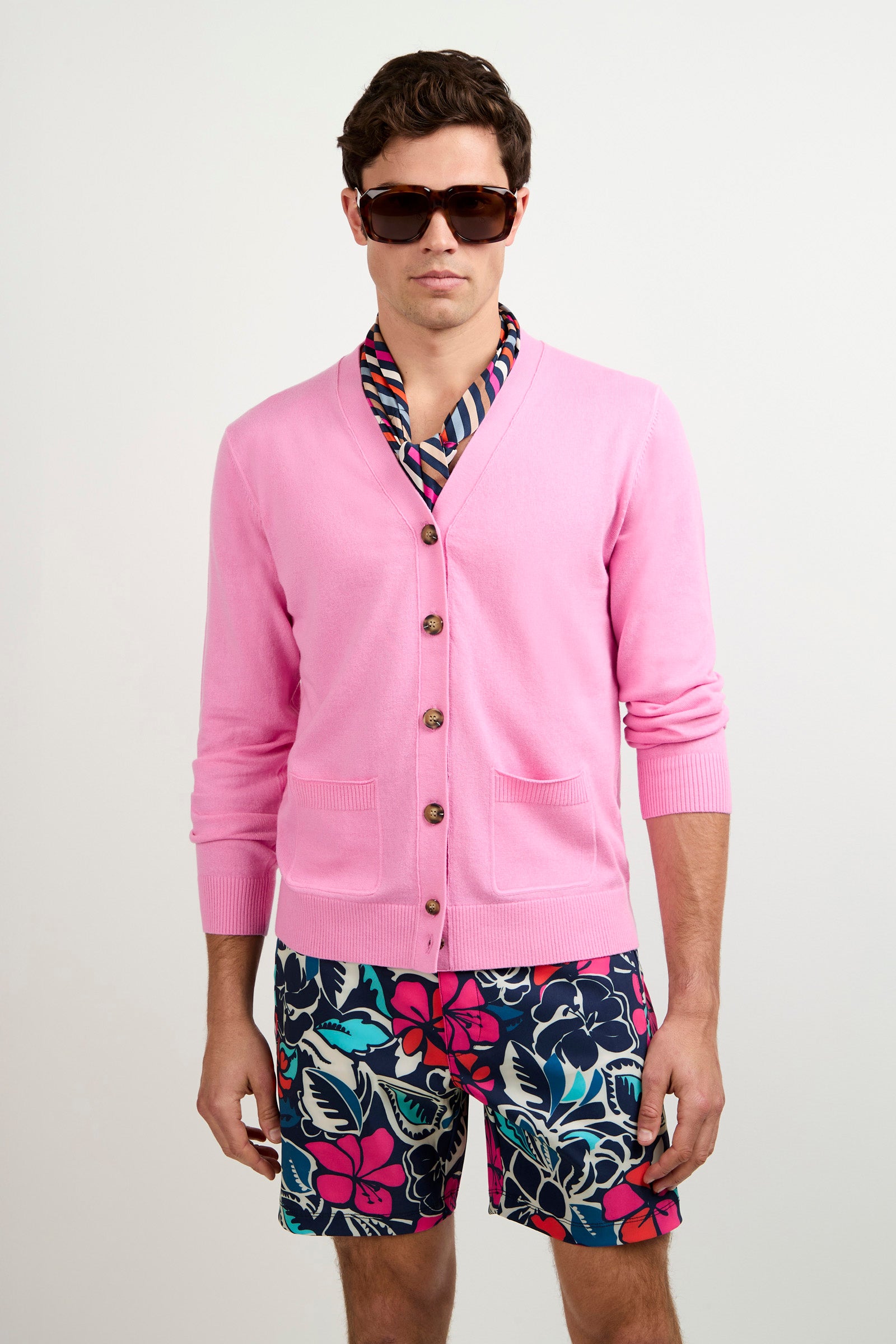 A man wearing sunglasses, the Jacques Cardigan in pink over a colorful patterned shirt, and navy blue shorts with a bright pink and teal floral print stands against a plain white background.