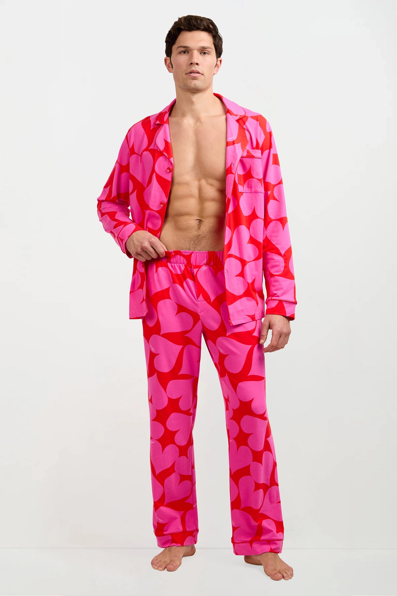 A man stands barefoot in the Men's Classic PJ Set - Hearts, featuring bright pink pajamas with large red hearts. The top is unbuttoned to reveal his bare chest. The background is plain white.