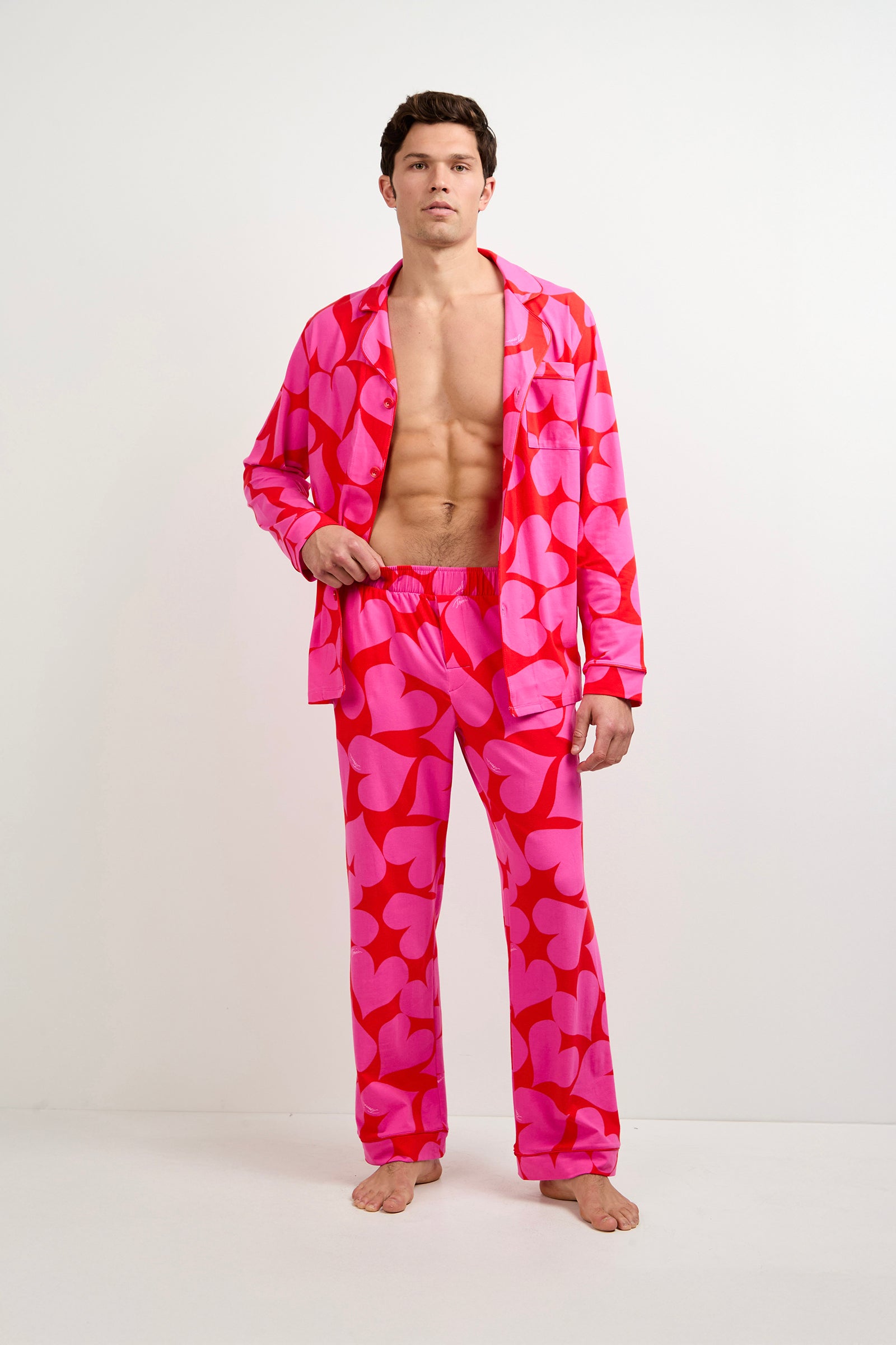 A man stands against a white background wearing the Men's Classic PJ Set - Hearts, featuring bright pink pajamas with bold red heart patterns and an unbuttoned shirt revealing his chest.