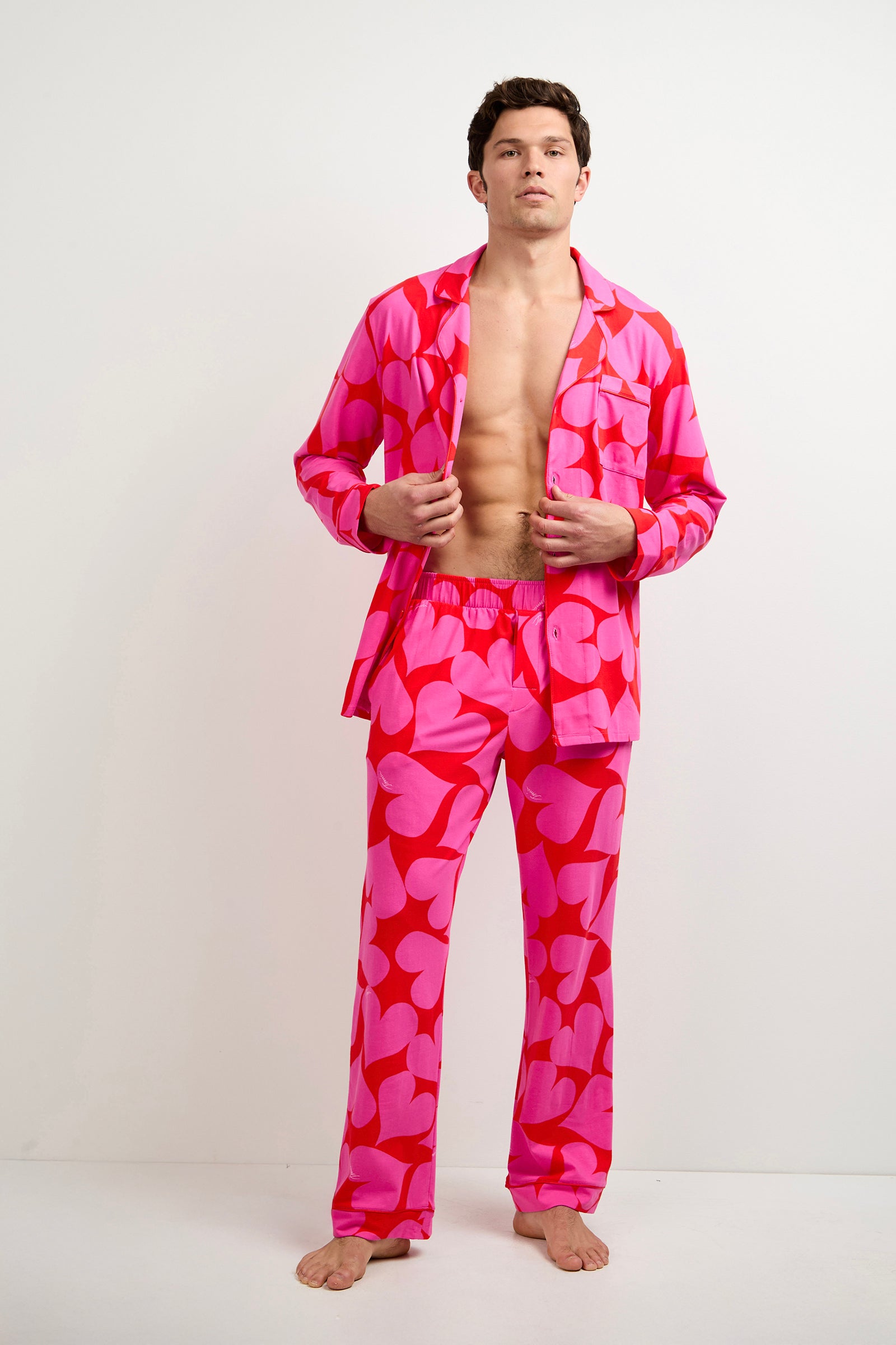 A man stands barefoot against a plain white background, wearing the Men's Classic PJ Set - Hearts, featuring vibrant pink fabric with bold red heart patterns and an open pajama shirt revealing his bare chest.