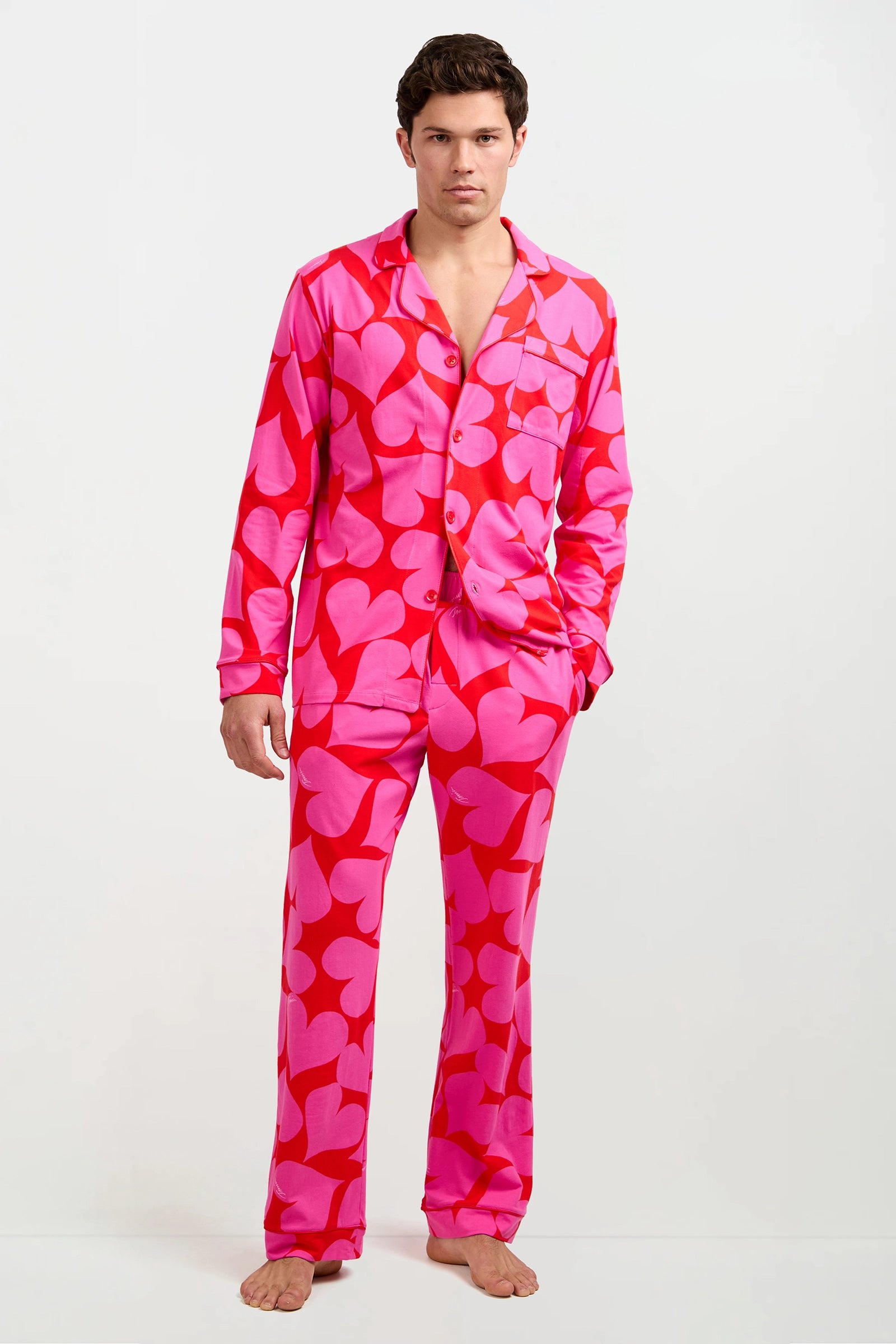 A person stands barefoot in the Men's Classic PJ Set - Hearts, featuring bright pink fabric with bold red heart patterns, long sleeves, a collar, and buttons, posing against a plain white background.