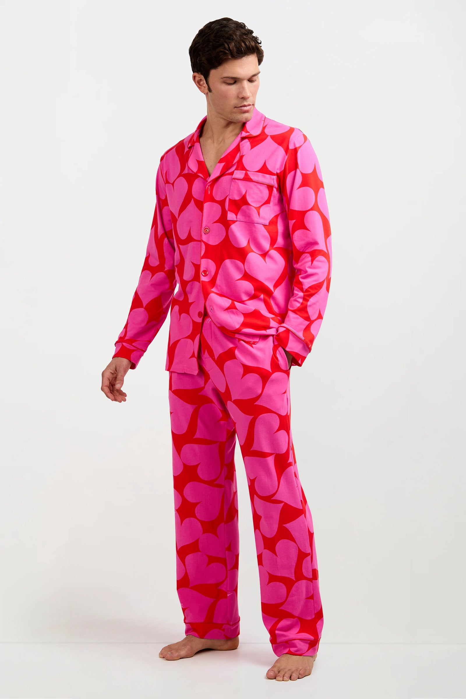 A man stands barefoot, looking down to his left, dressed in the Men's Classic PJ Set - Hearts: a bright pink and red heart-patterned long-sleeve button-up shirt with matching pants. The background is plain white.