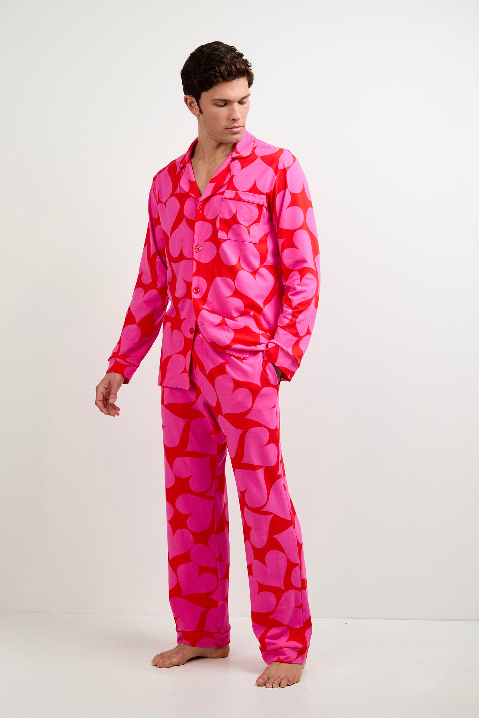 A person stands barefoot against a plain backdrop, wearing the Men's Classic PJ Set - Hearts featuring vibrant pink fabric with bold red abstract heart patterns, and looks slightly to the side with one hand lowered.
