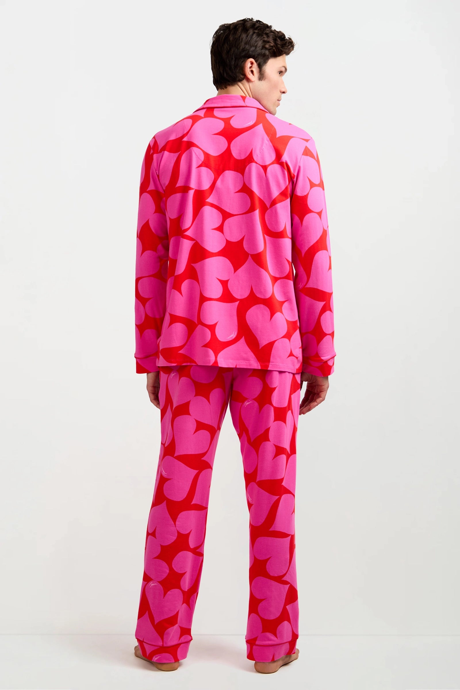 A person faces away, wearing the Men's Classic PJ Set - Hearts in bright pink and red with a bold heart pattern. The background is plain white.