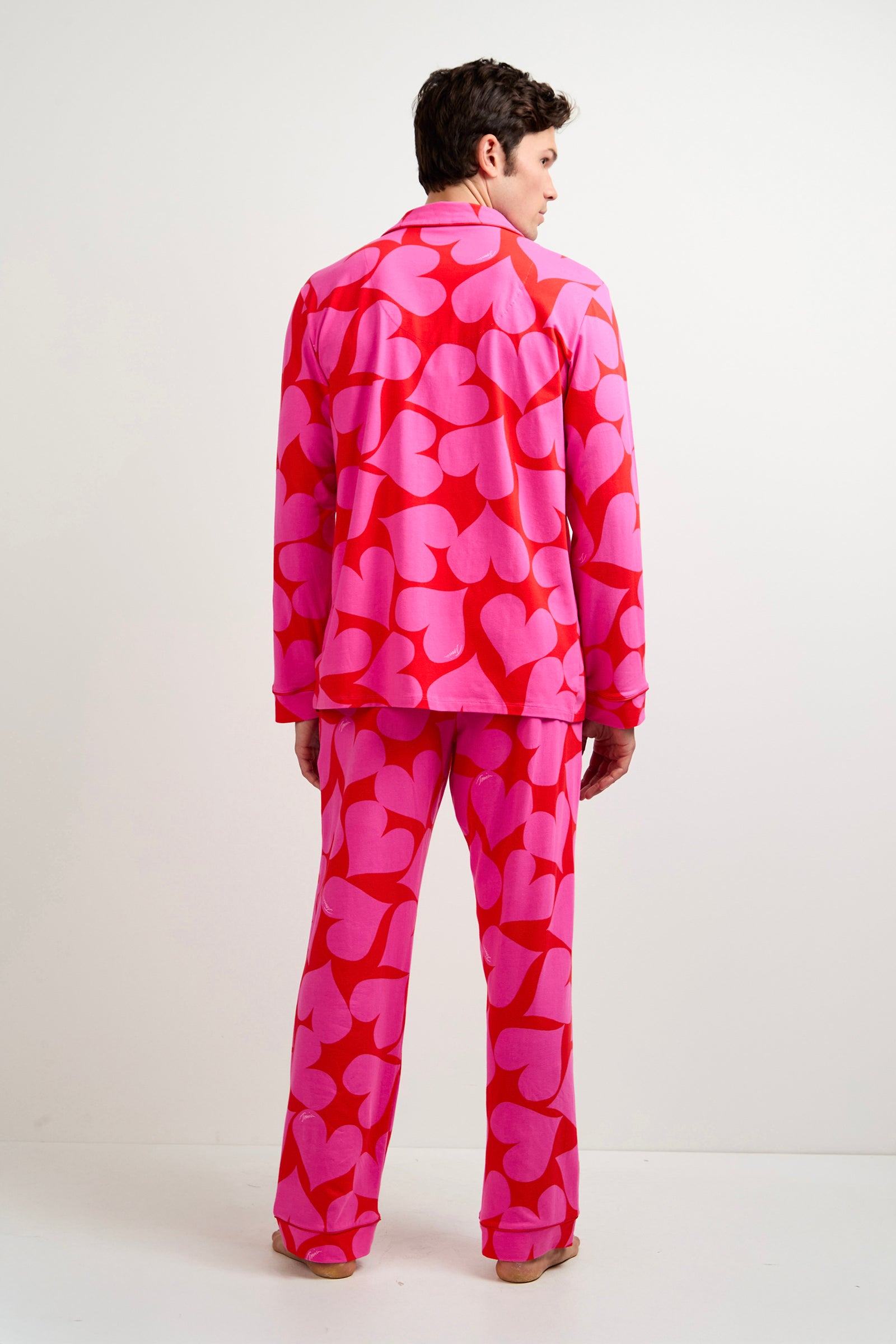 A man faces away, wearing the Men's Classic PJ Set - Hearts, a bright pink pajama set with large red heart patterns, against a plain white background.