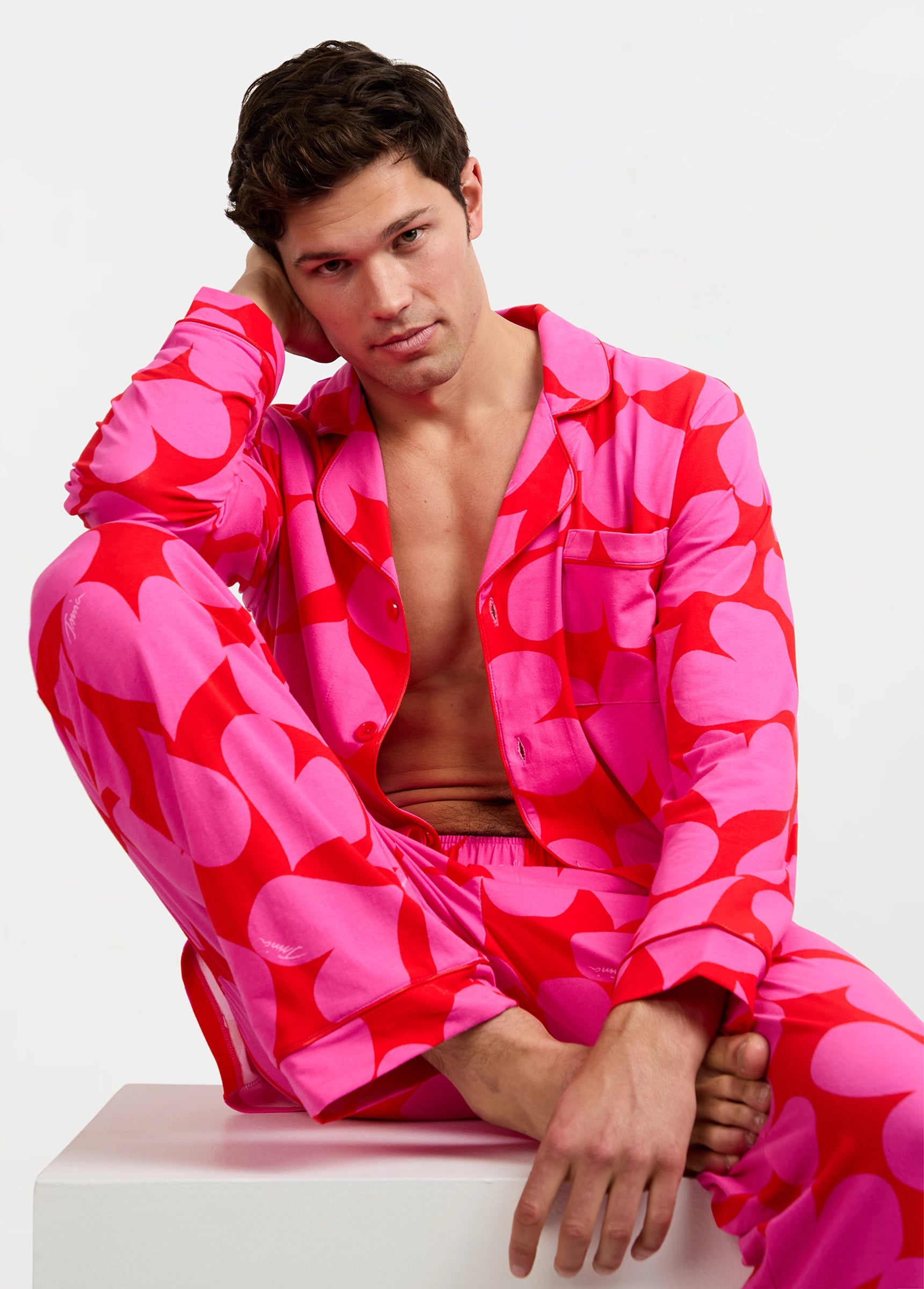 A man wears the Men's Classic PJ Set - Hearts, featuring bright pink fabric with large red hearts. Sitting on a white block, he leans his head on one hand and gazes at the camera, his shirt partially unbuttoned.