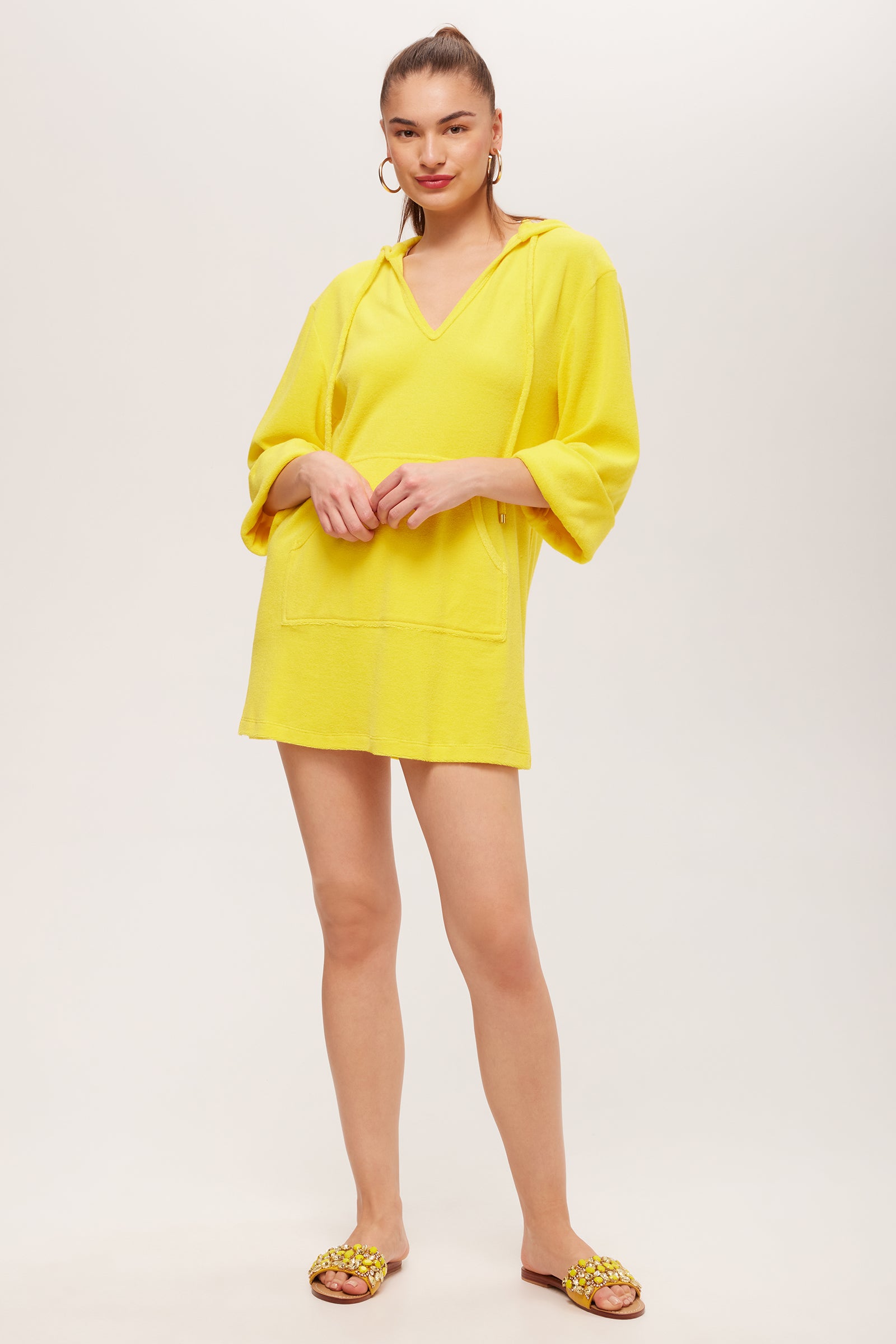 A woman stands against a white background, wearing the Poolside 3/4-Sleeve Hoodie in Desert Sun, a mini dress with three-quarter sleeves and front pockets. Her hair is tied back, and she pairs it with hoop earrings and embellished brown slide sandals.