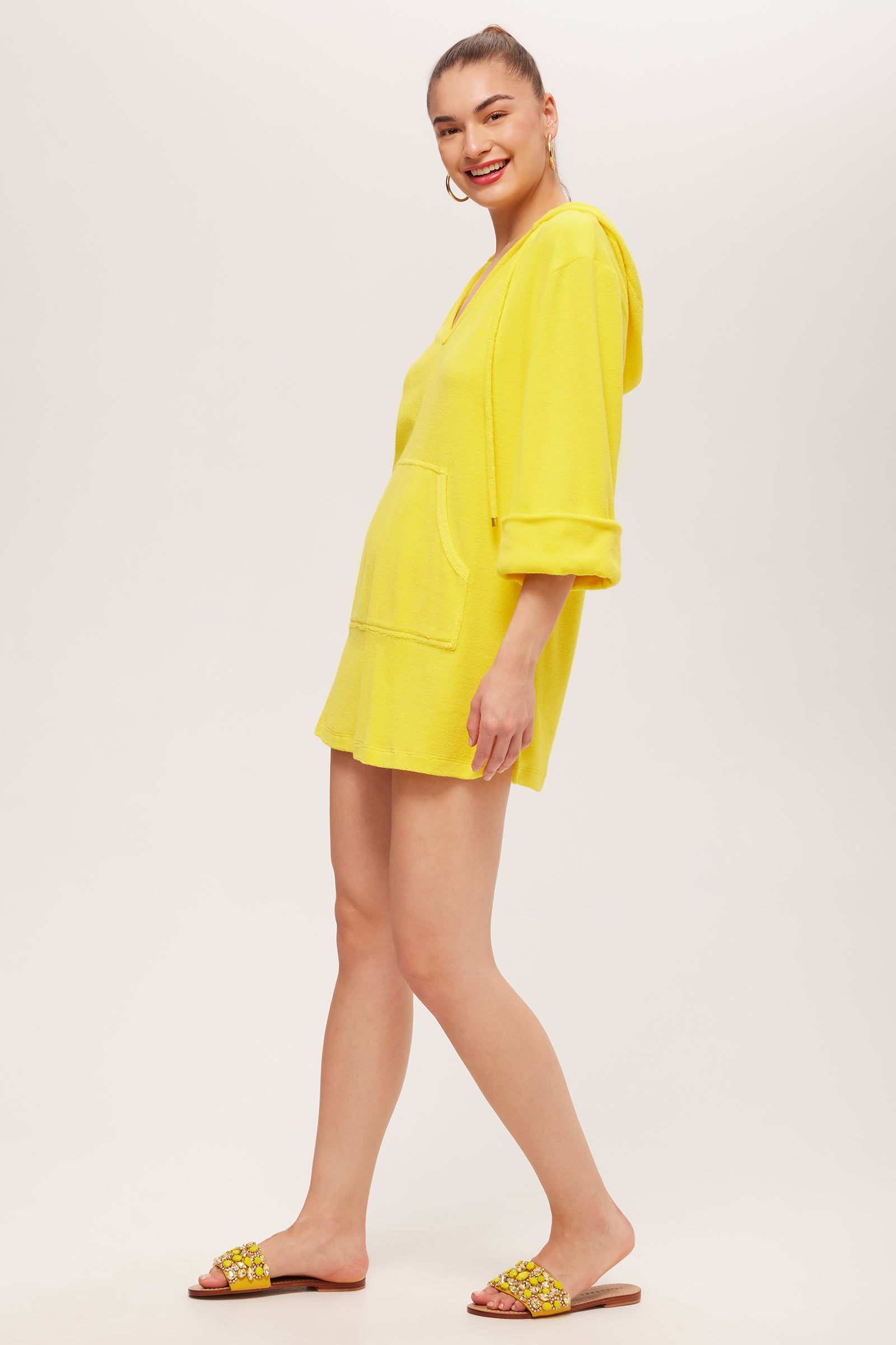 A woman smiles, wearing the Poolside 3/4-Sleeve Hoodie - Poolside Terry in bright yellow with rolled sleeves and a front pocket, paired with yellow embellished slide sandals. Her hair is tied back as she poses against a light background.