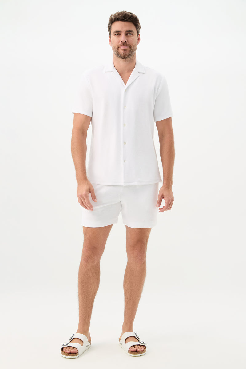 A man with short brown hair and a trimmed beard stands against a plain white background, wearing the Felix Short-Sleeve Knit Shirt - Linsk Terry, paired with matching white shorts and white sandals.
