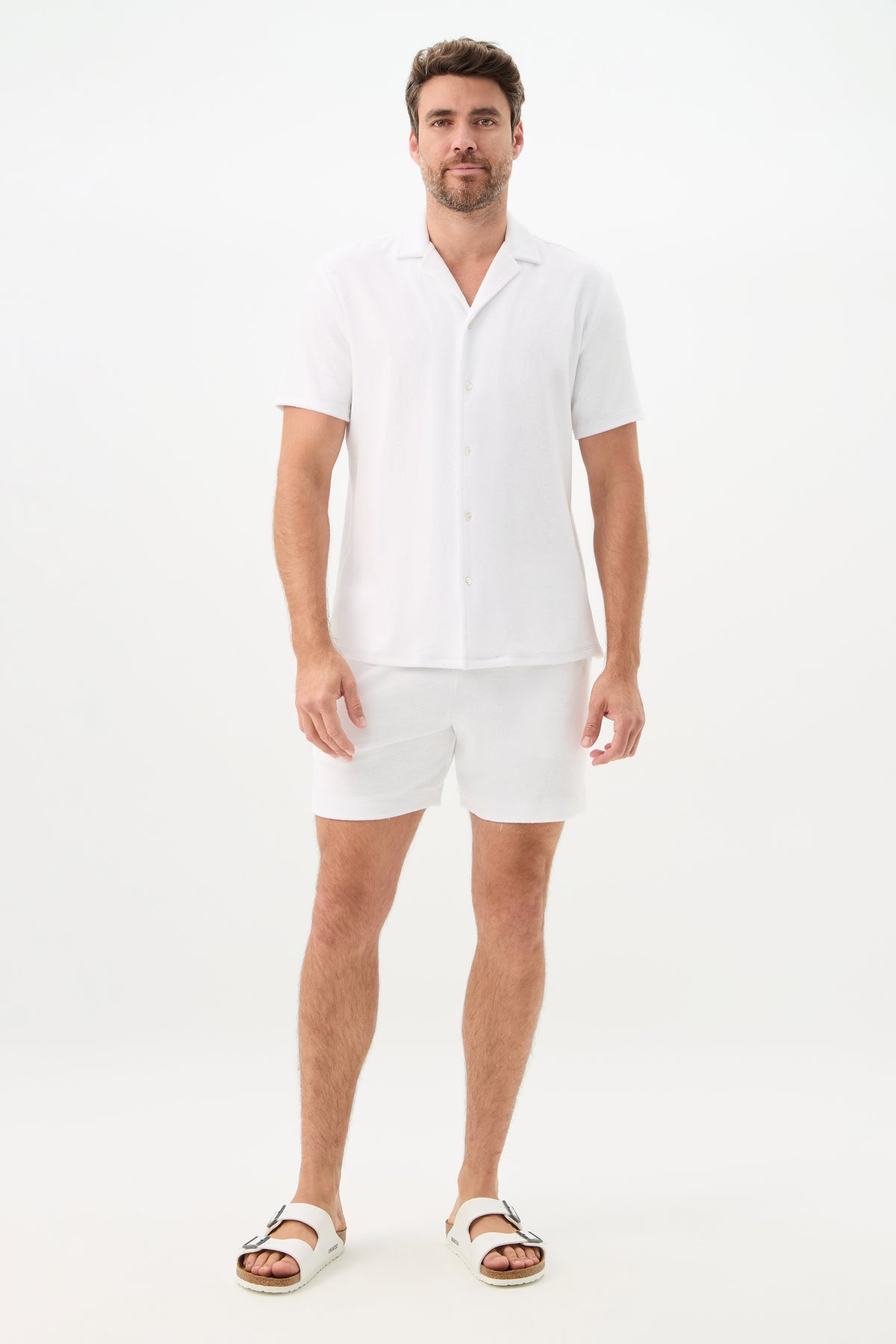A man stands against a plain white background wearing a white short-sleeve button-up shirt, Lounge Knit Short - Linsk Terry in white, and white sandals.