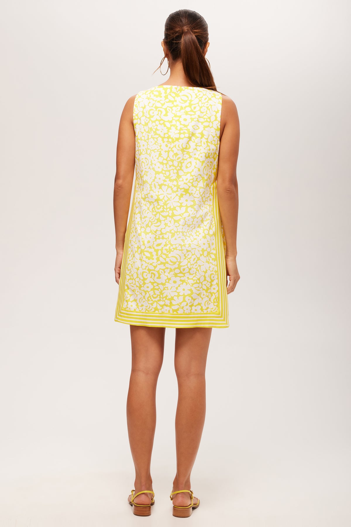 A woman with a ponytail faces away, wearing the Brynne 3 Sleeveless Dress in Poolside Gossip—a short yellow and white patterned dress with a square neckline—paired with flat sandals against a plain background.