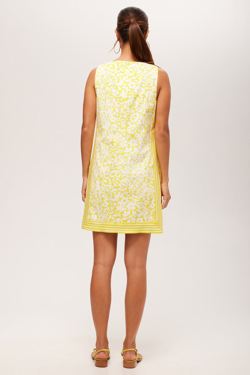 A woman with a ponytail faces away, wearing the Brynne 3 Sleeveless Dress in Poolside Gossip—a short yellow and white patterned dress with a square neckline—paired with flat sandals against a plain background.