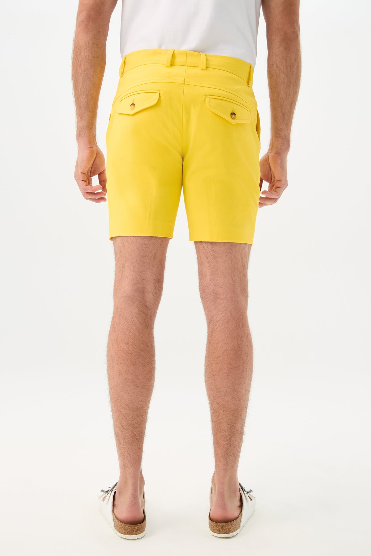 A man faces away from the camera, wearing a white t-shirt, Cyrus Short - Sunset Stretch in bright yellow with belted waist and buttoned back pockets, and striped slip-on shoes, set against a plain white background.