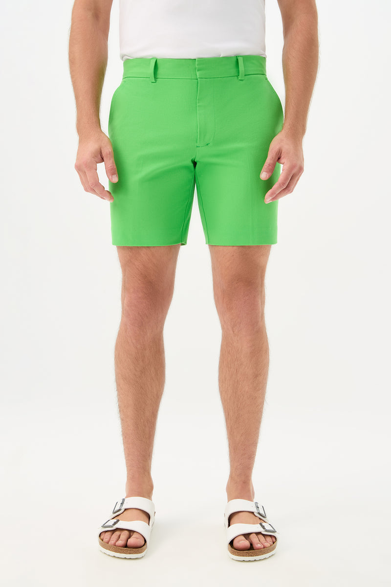 A person is shown from the waist down, wearing the Cyrus Short - Cotton Bi-Stretch in vert with a white shirt and white sandals, standing against a plain white background.