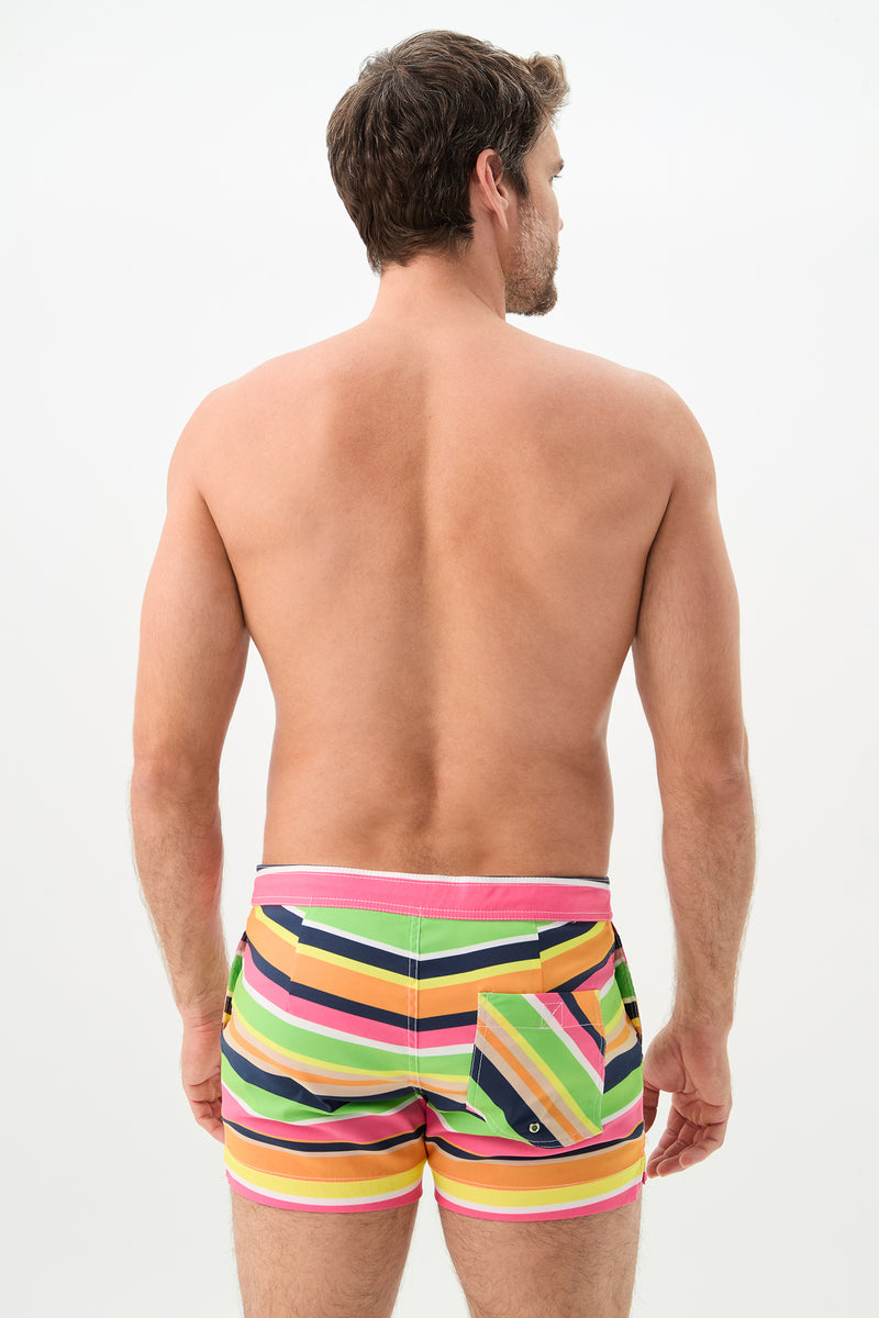 A man faces away from the camera, modeling the Chico Swim Trunk - Splash Stripe, which features colorful diagonal stripes in pink, yellow, green, orange, and black against a plain white background.