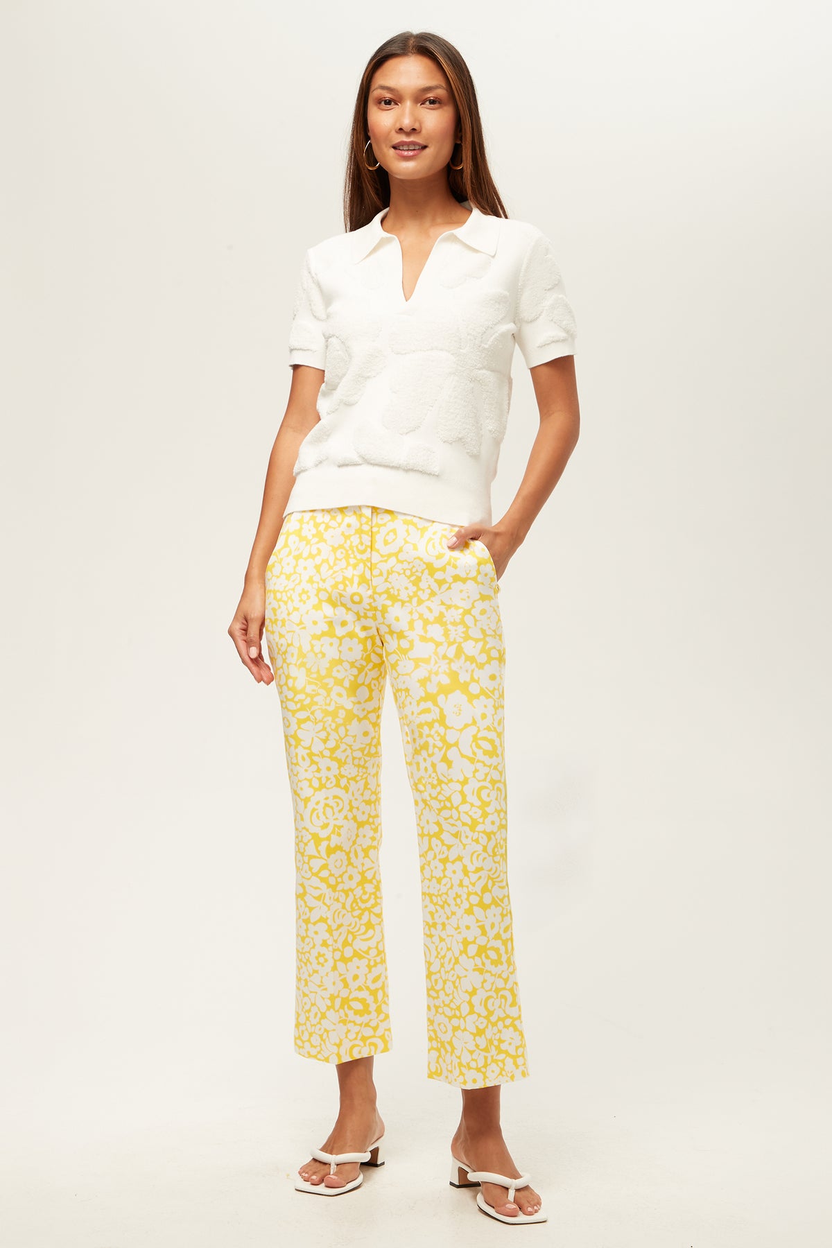 A woman smiles, wearing the Lulu Cropped Pant - Poolside Gossip with a textured white short-sleeve top and white sandals. She has long straight hair, hands in her pockets, and stands against a plain white background.