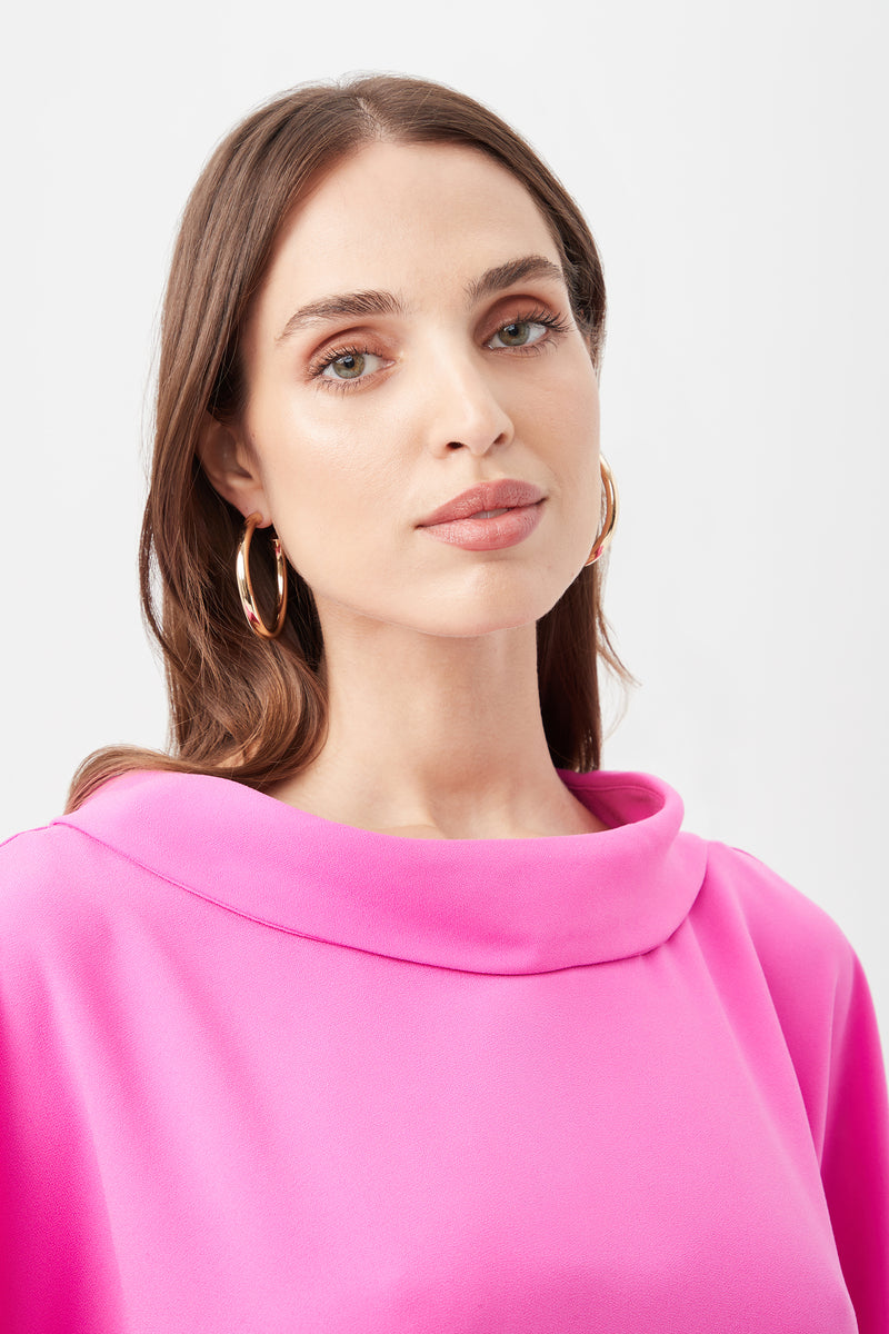 A woman with light eyes and long brown hair wears gold hoop earrings and the Honeysuckle Boatneck Dolman-Sleeve Top, featuring a wide collar, as she stands against a plain white background.