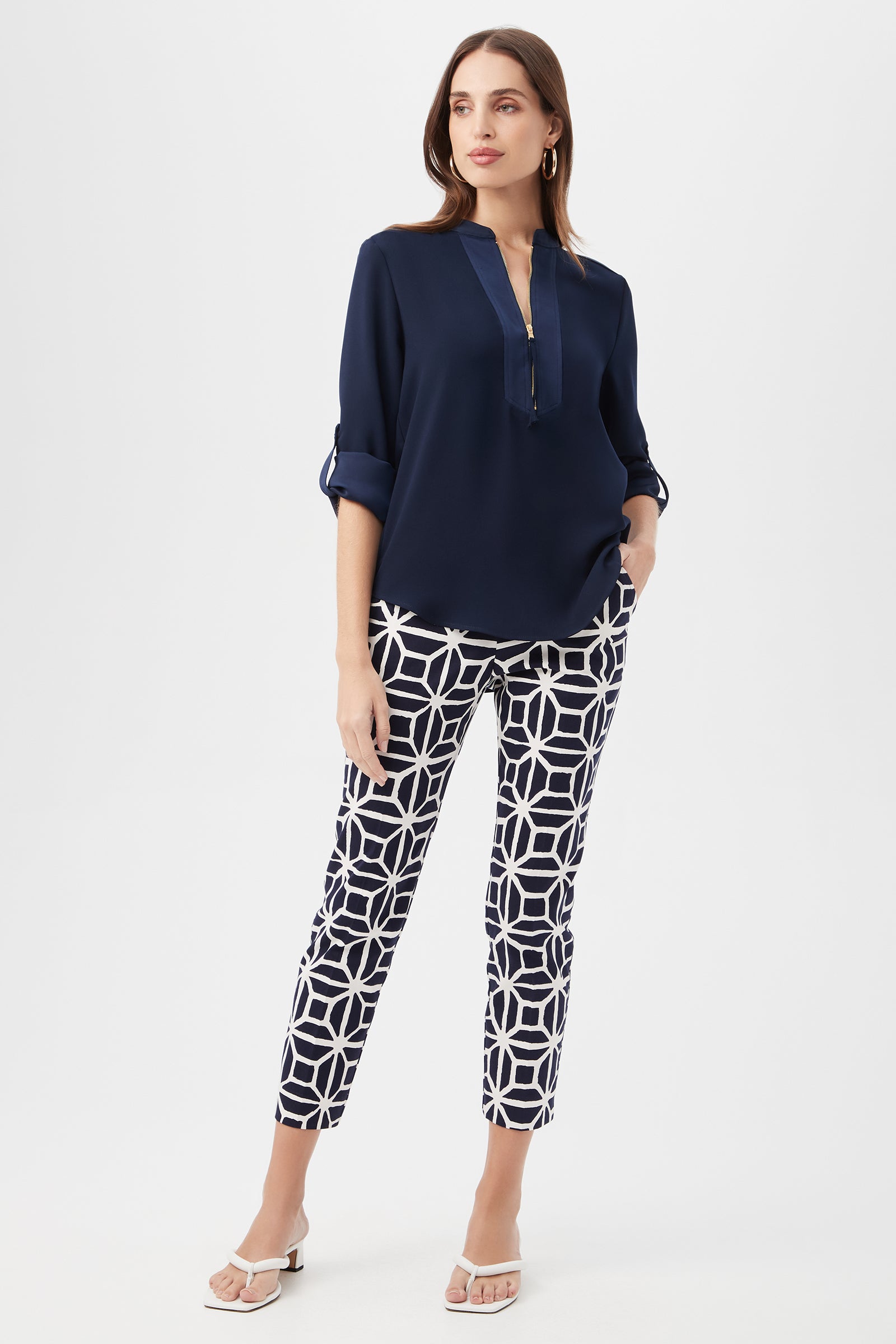 A woman wears the Kaiko 3/4-Sleeve Half-Zip Top with a V-neck and rolled sleeves, paired with white ankle-length pants featuring a bold navy geometric pattern. She completes her look with white heeled sandals and hoop earrings.