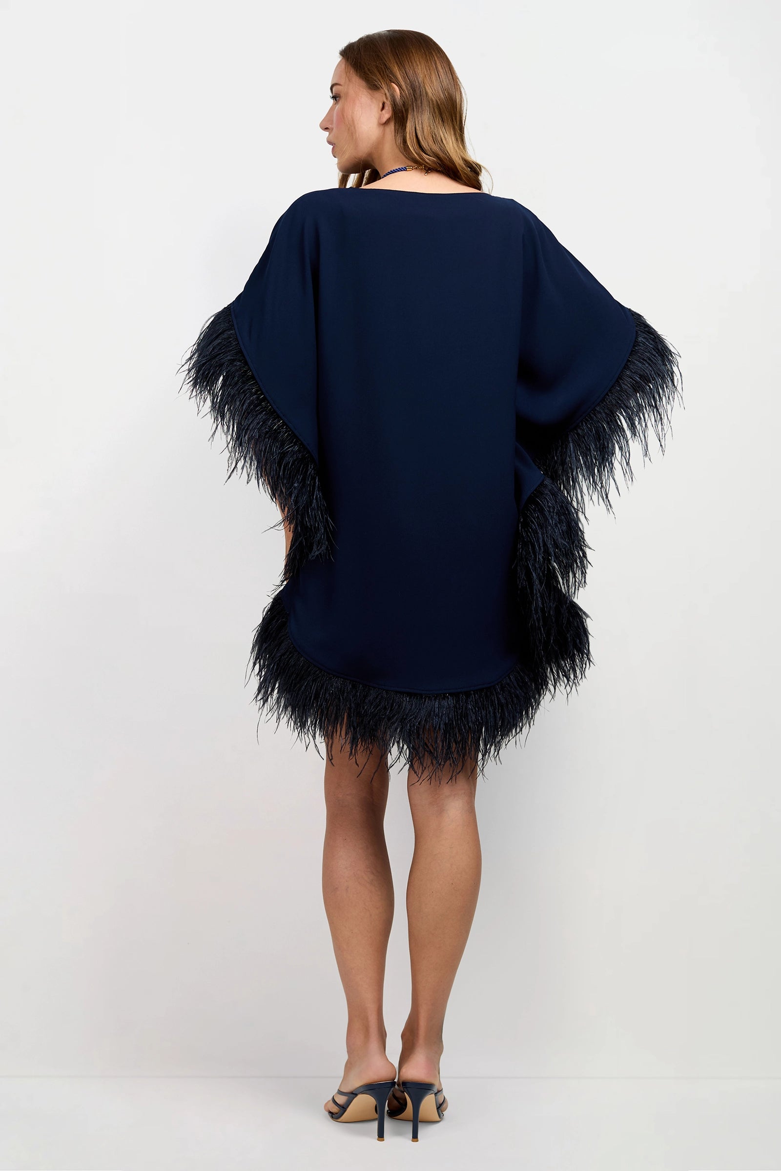A woman faces away, wearing the Mini Neena Caftan Dress—a loose navy blue dress with black feather trim on the sleeves and hem—and black heeled sandals, set against a plain white background.