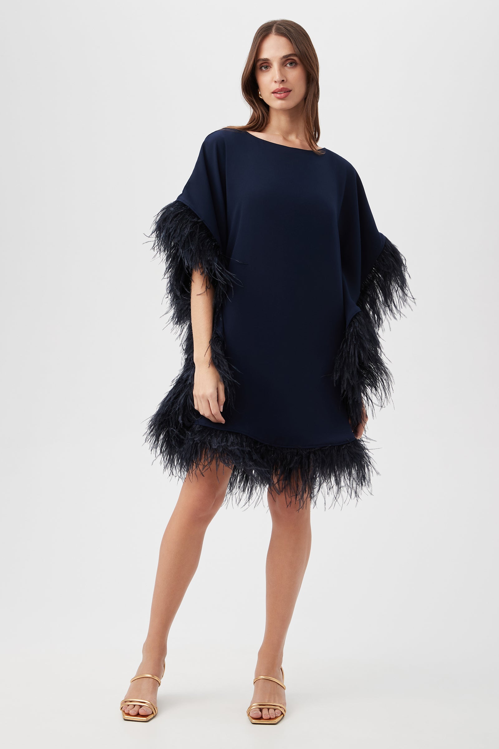 A woman models the Mini Neena Caftan Dress in indigo, featuring black feather trim on the sleeves and hem. She pairs it with strappy gold sandals and stands against a plain white background.