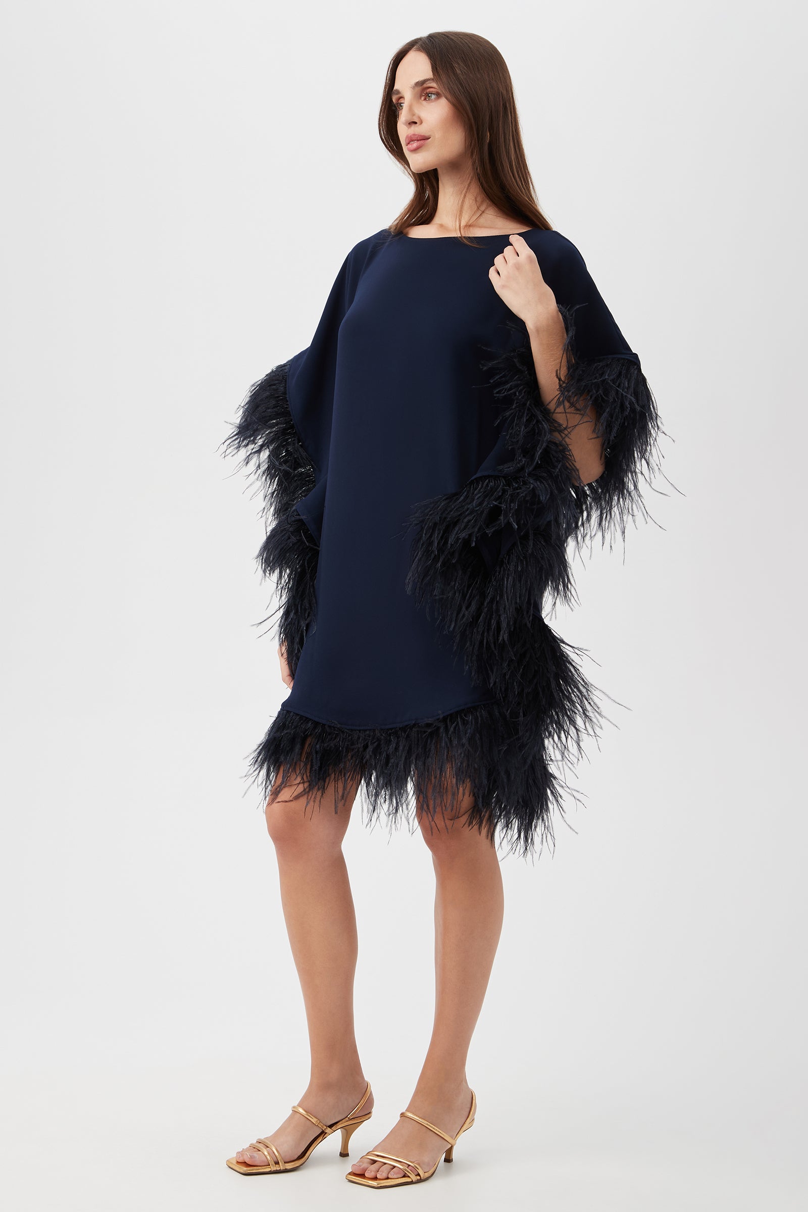 A woman wears the Mini Neena Caftan Dress, a loose-fitting navy blue dress with black feather trim on the sleeves and hem, paired with gold heeled sandals, standing against a plain white background.