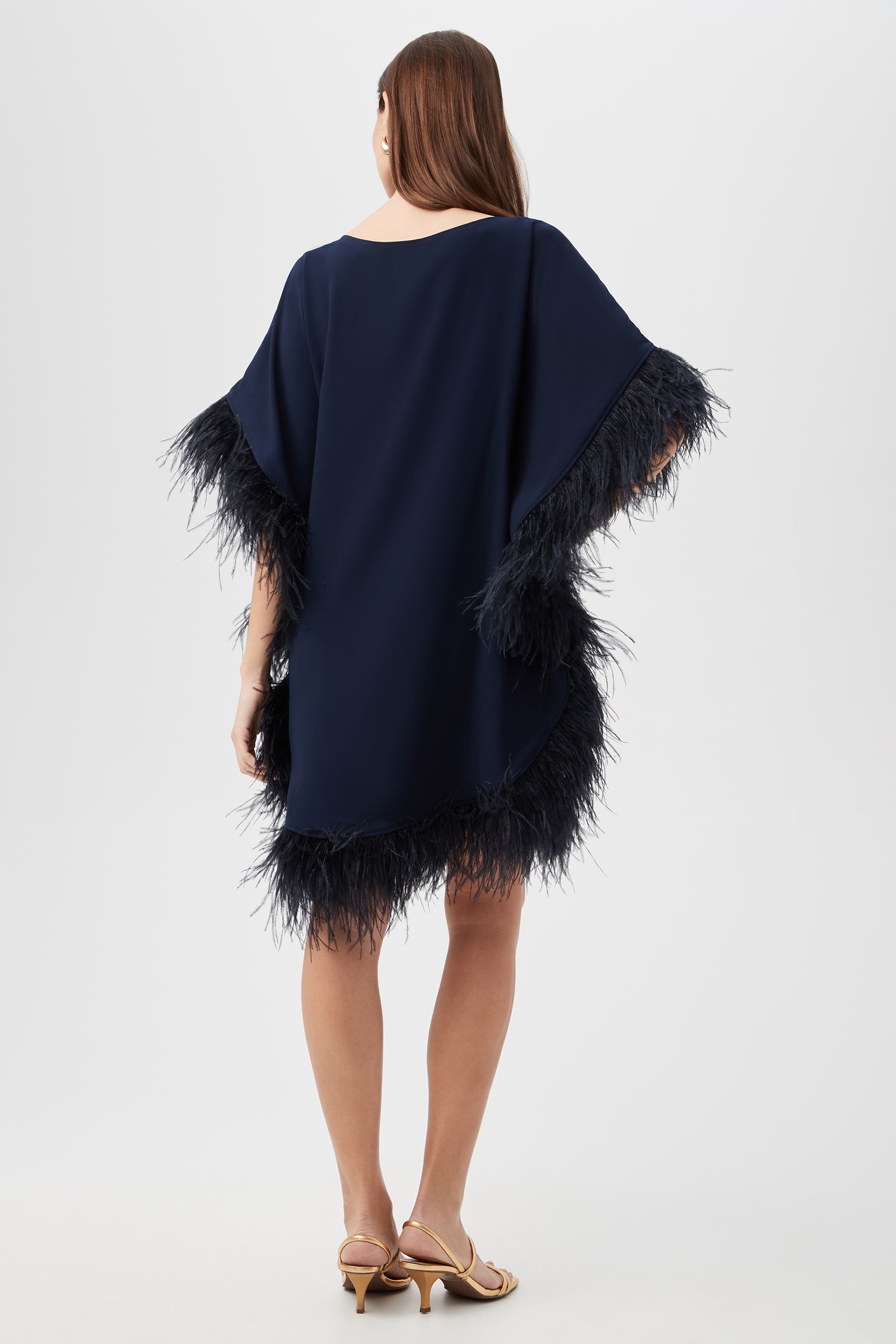 A woman faces away, wearing the Mini Neena Caftan Dress in navy with feather-trimmed sleeves and hem. She has straight brown hair and beige heeled sandals, set against a light background.
