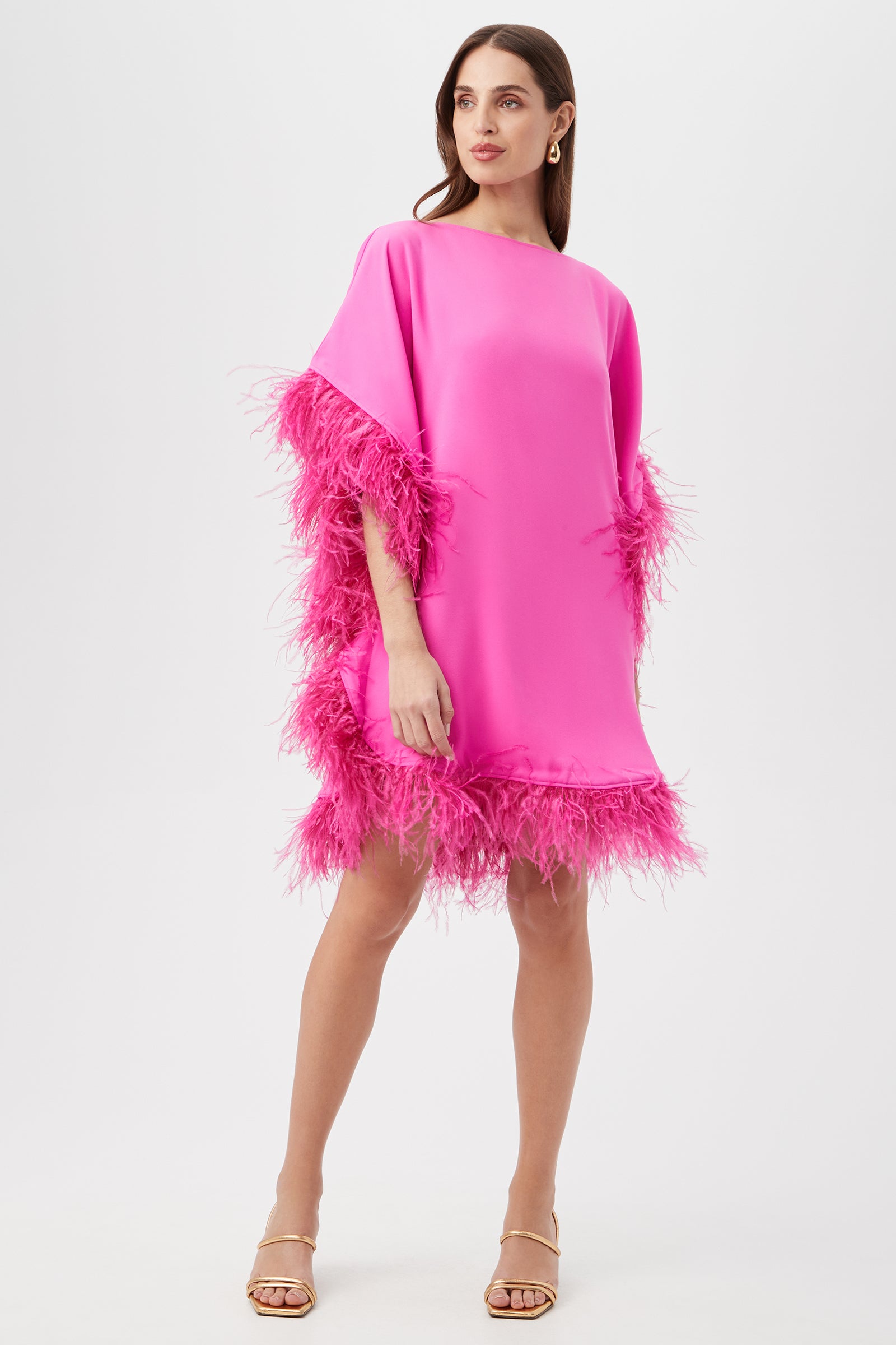 A woman in the Mini Neena Caftan Dress in Snapdragon, featuring loose fit and matching feather trim on the sleeves and hem, paired with nude heeled sandals, stands against a plain white background.