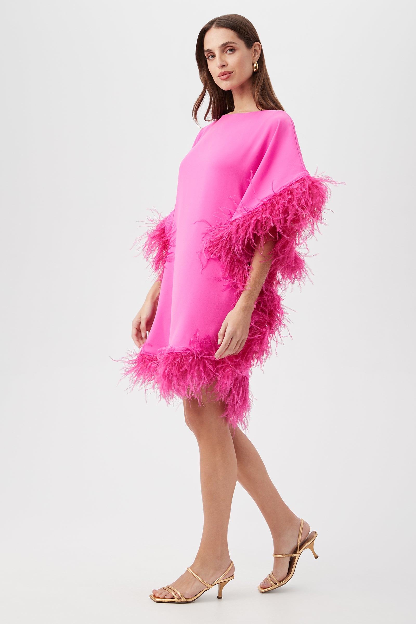 A woman wears the Mini Neena Caftan Dress, a bright pink, loose-fitting dress with feather trim on the sleeves and hem. She pairs it with gold heeled sandals and stands against a plain white background.