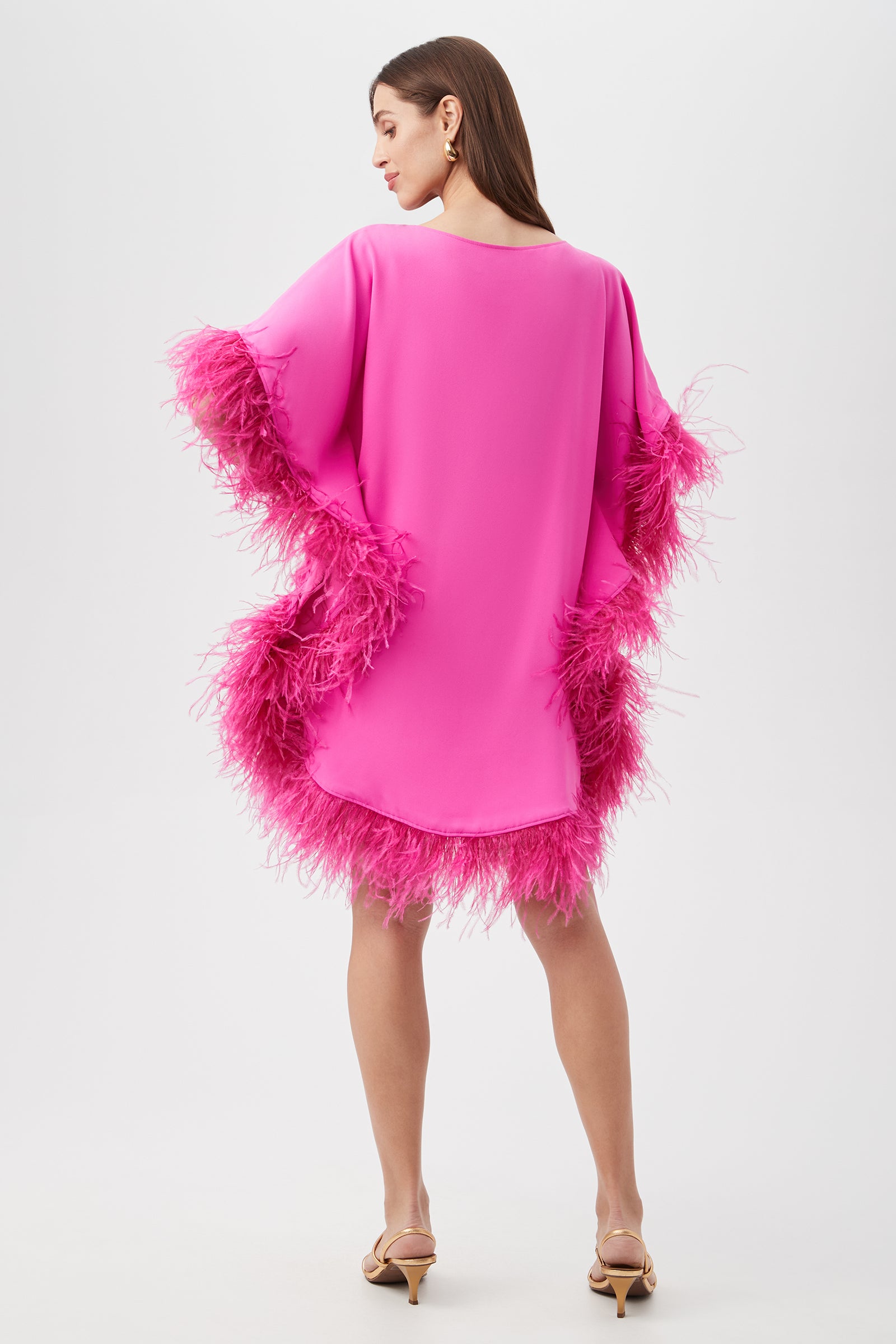 A woman with straight brown hair faces away, modeling the Mini Neena Caftan Dress—a loose, bright pink design with feathered trim at the sleeves and hem—paired with strappy nude heels against a plain white background.