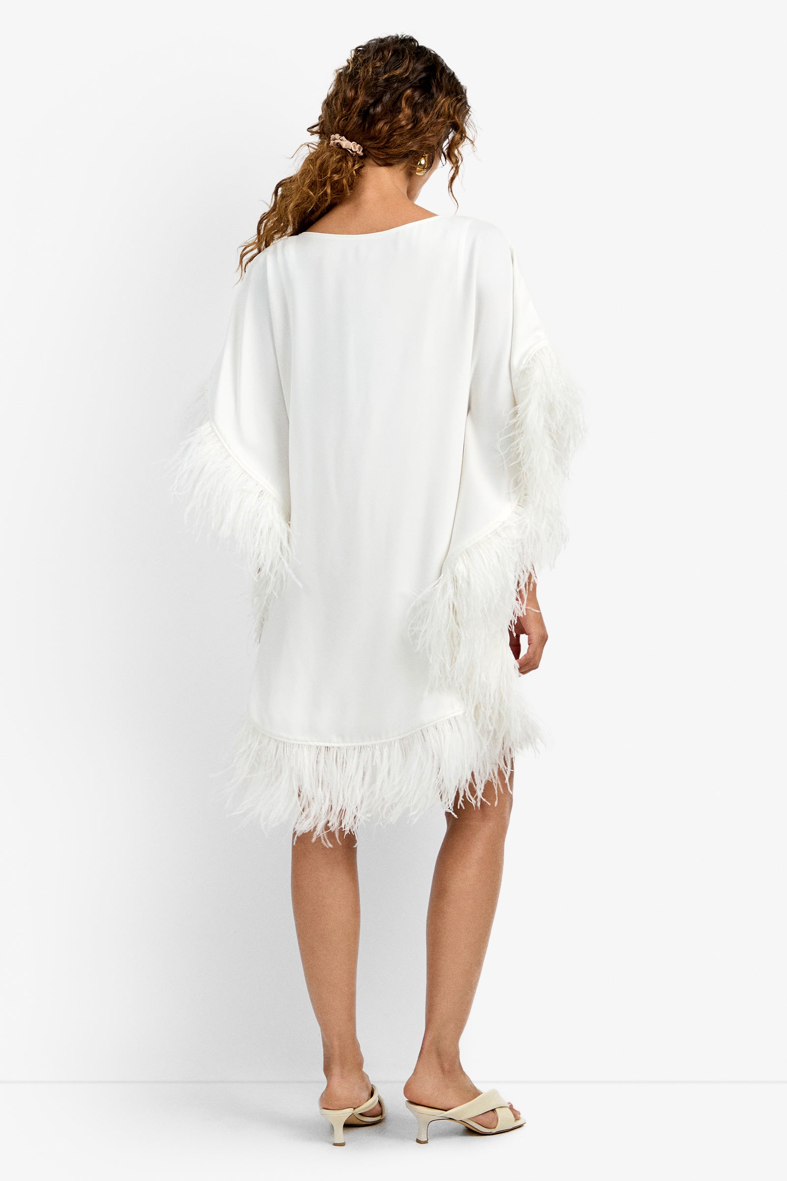 A woman with curly hair stands facing away against a plain white background, wearing the Mini Neena Caftan Dress—a loose white dress featuring feather trim on the sleeves and hem—paired with beige heeled sandals.