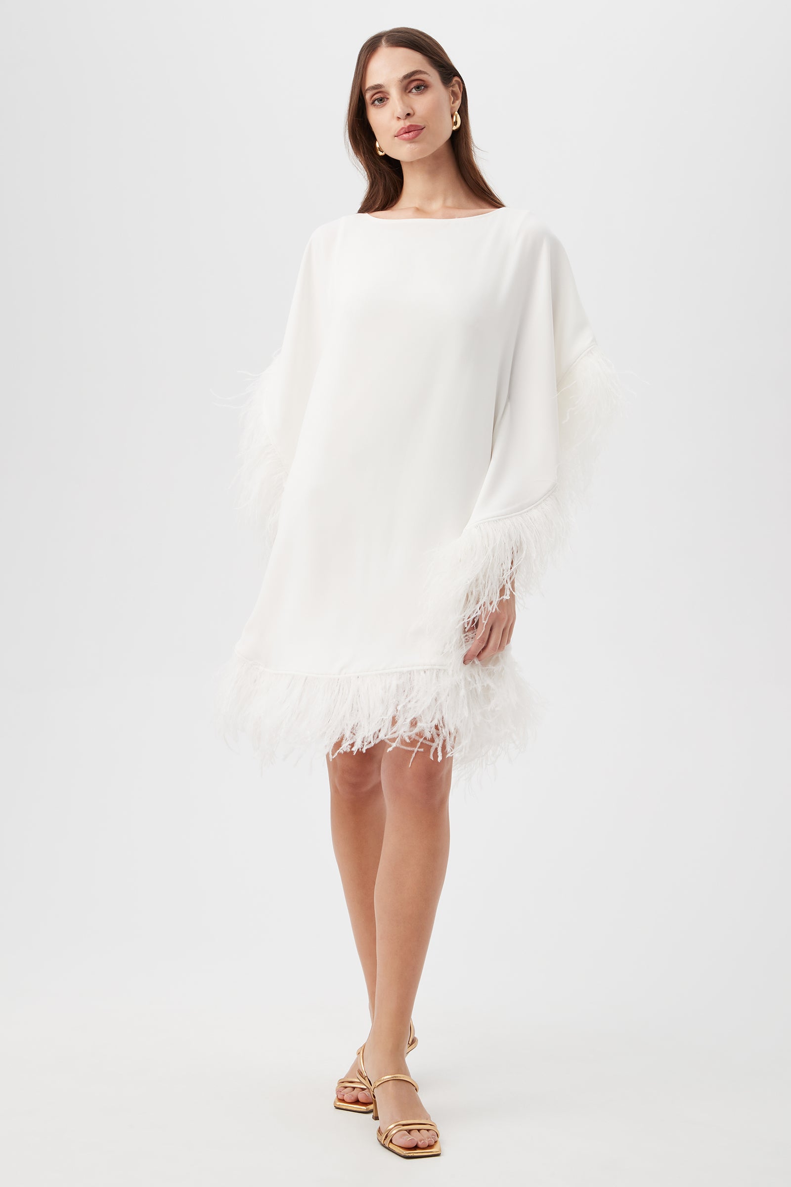 A woman stands against a white background wearing the Mini Neena Caftan Dress in Whitewash, featuring feather trim on the sleeves and hem. She accessorizes with gold hoop earrings, gold sandals, and has long brown hair.