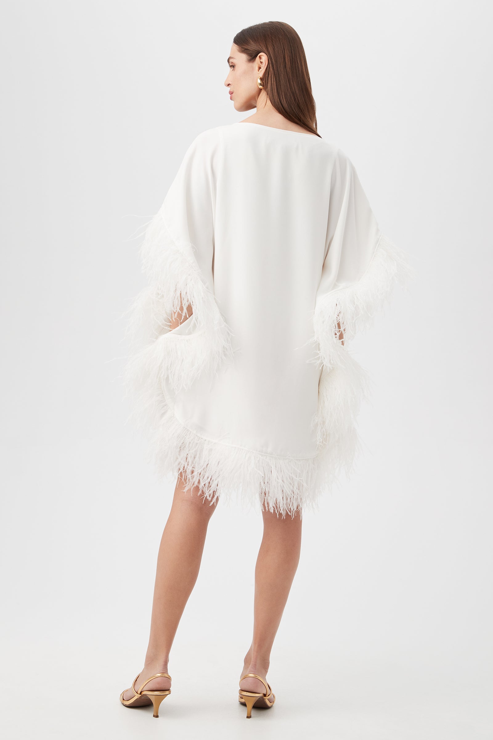 A woman with straight brown hair faces away, wearing the Mini Neena Caftan Dress—a loose white dress with feather-trimmed sleeves and hem—paired with tan heeled sandals, set against a plain white background.