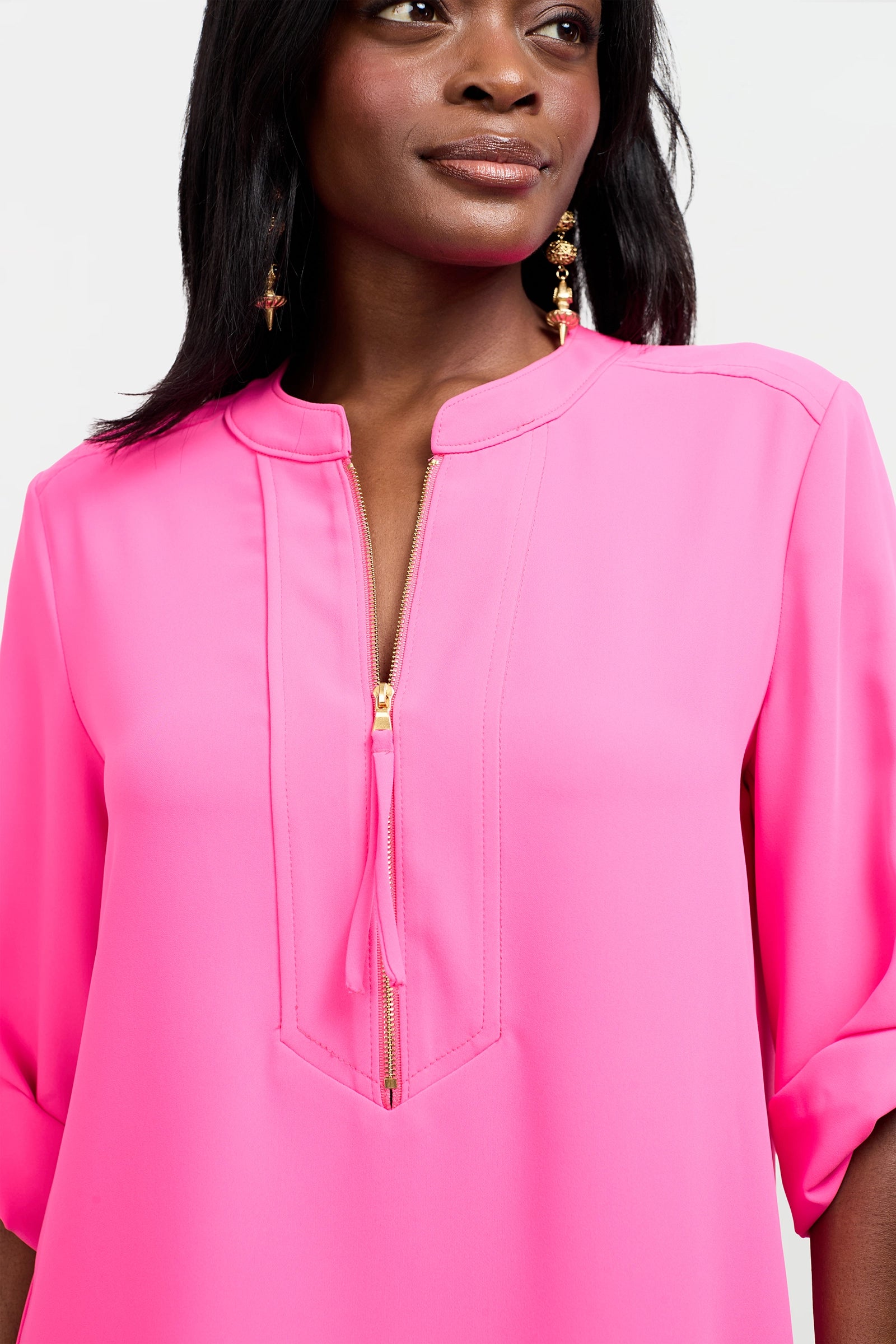 A woman in the Kaiko 2 Dress, featuring a bright pink hue, gold front zipper, and 3/4 rolled sleeves, paired with gold statement earrings. Photo cropped above her elbows and just below her eyes.
