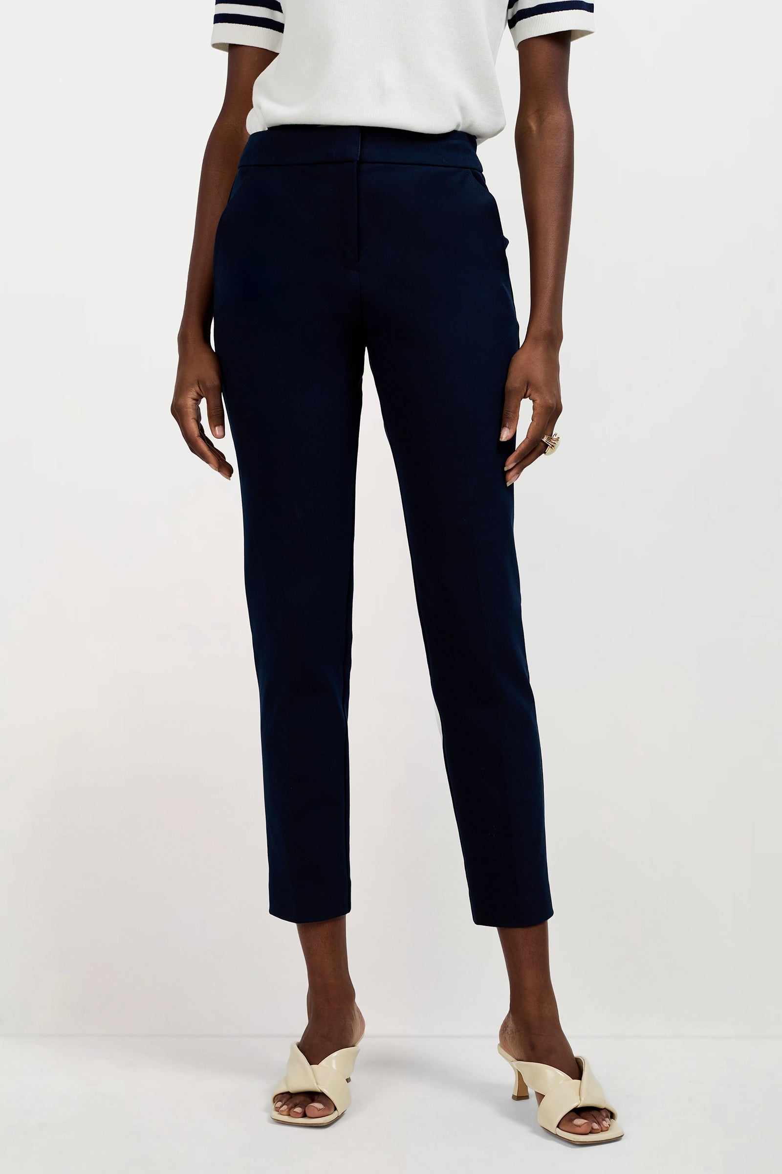 Wearing Aubree 2 Cigarette Pants in dark navy, a short-sleeved white top with dark striped cuffs, and cream open-toe heeled sandals, a person stands against a plain light background.