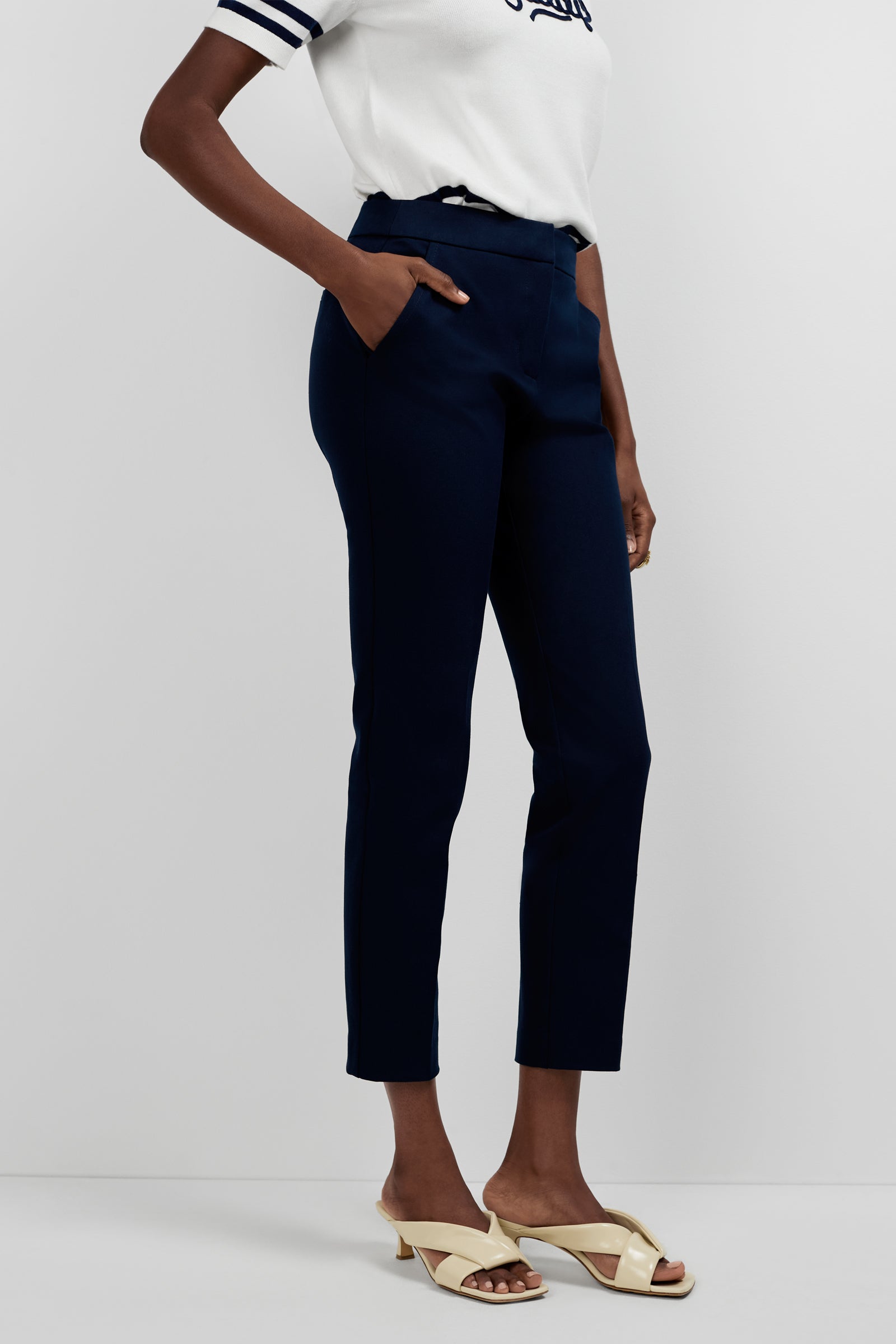 Wearing a white short-sleeve shirt with navy stripes, Aubree 2 Cigarette Pants in navy blue, and cream heeled sandals, a person stands with one hand in a pocket against a plain background.