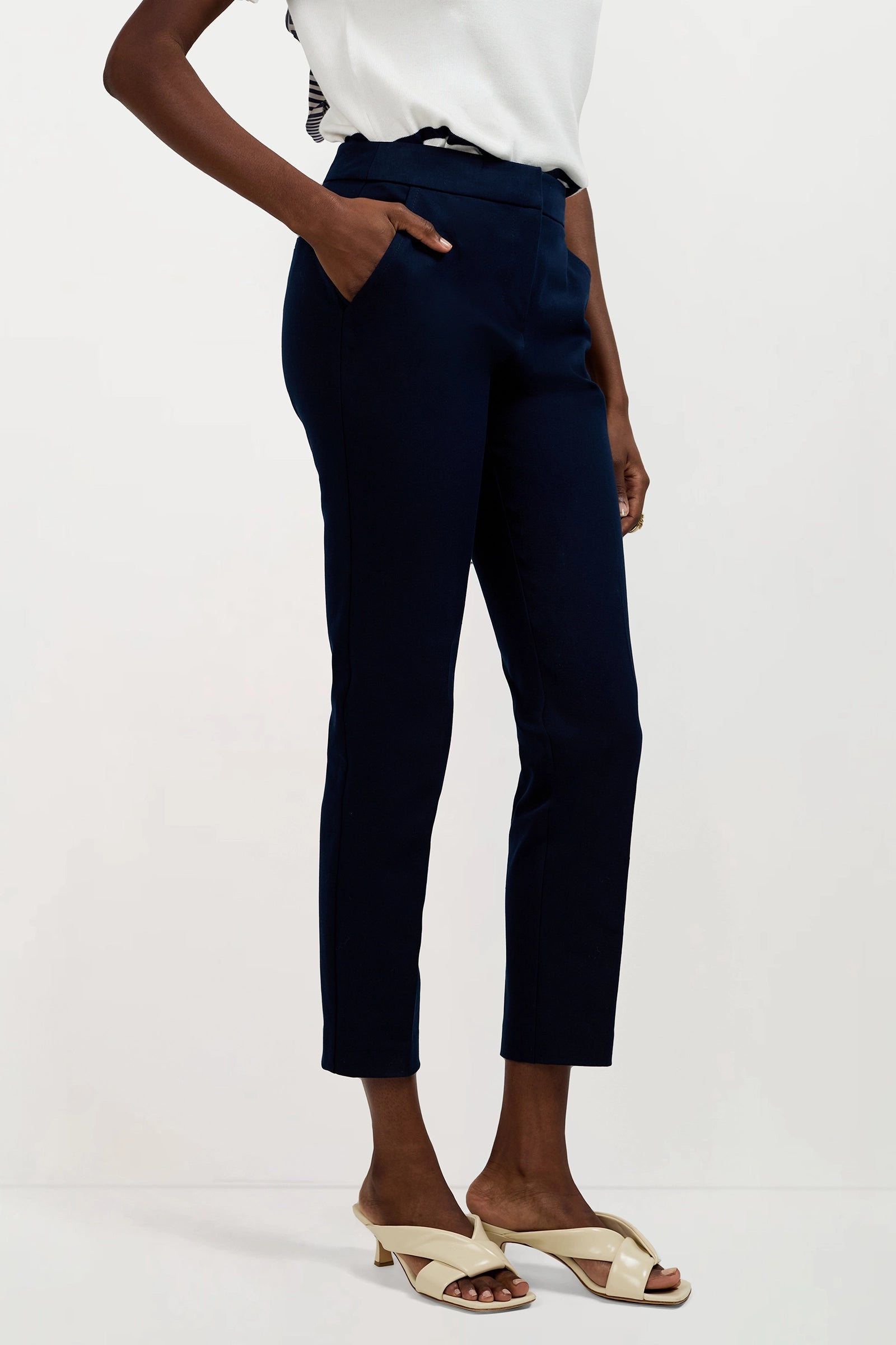 A person stands against a plain background wearing Aubree 2 Cigarette Pants in navy blue, paired with a white top and cream heeled sandals, with one hand in their pocket.