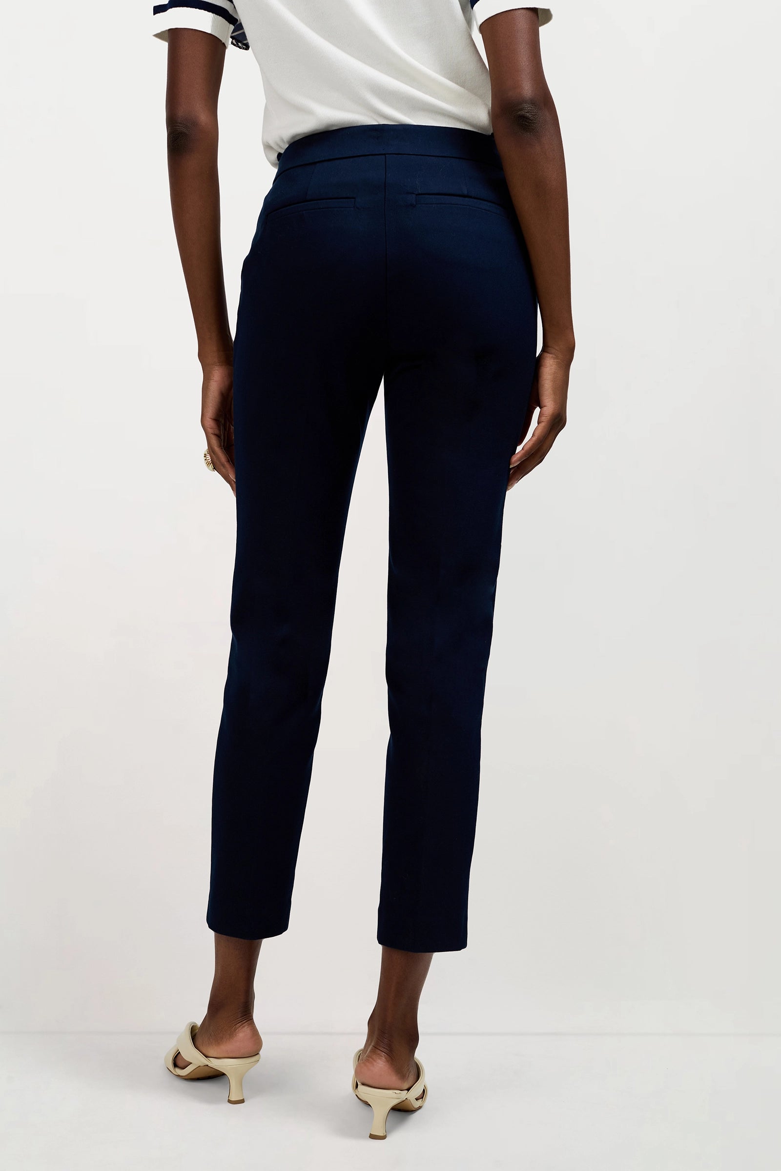 A person is shown from the back against a plain background, wearing Aubree 2 Cigarette Pants in dark navy, a short-sleeve white top, and cream-colored heeled sandals.