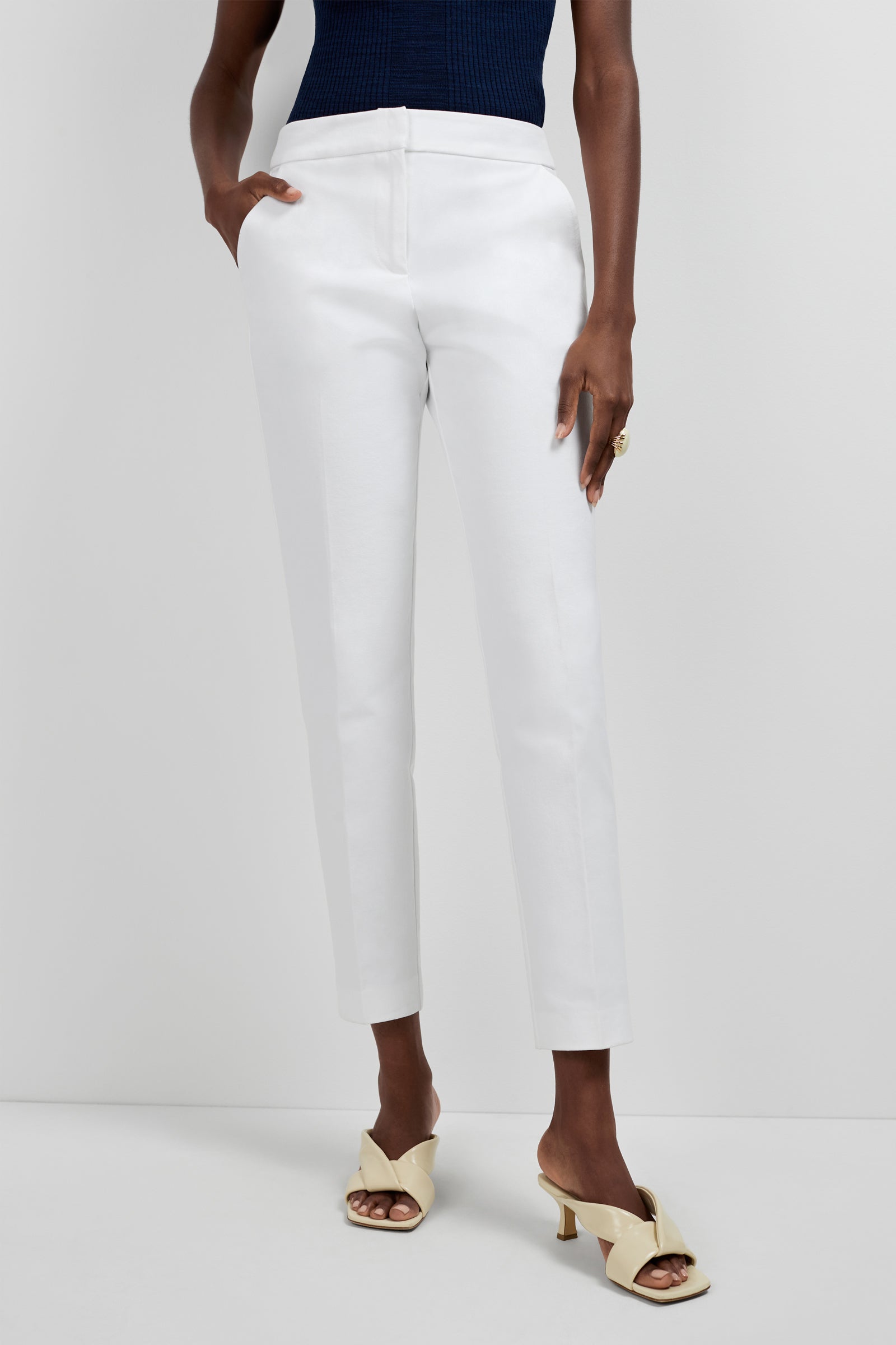 A person stands against a white background, wearing Aubree 2 Cigarette Pants in white, a navy sleeveless top, and cream open-toe heeled sandals, with one hand in their pocket.